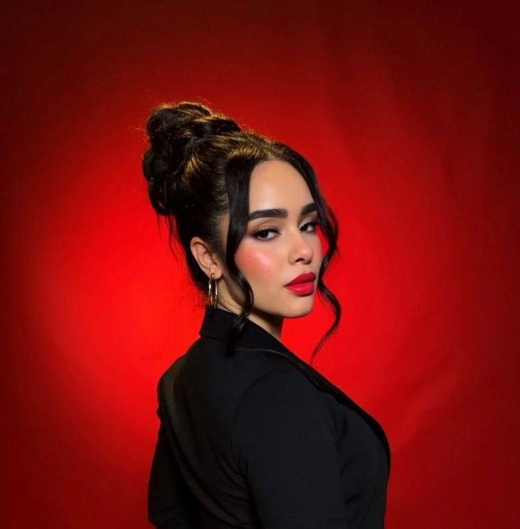 Young woman with dark hair in an updo, wearing a black top, gold hoop earrings, and red lipstick, against a red background.