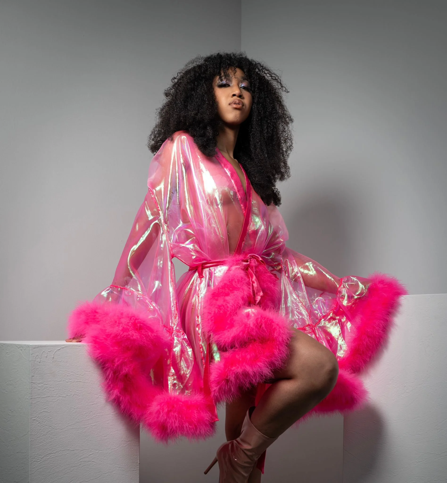 A woman with curly black hair wearing a pink, iridescent, transparent coat with pink feather trim sitting on a white ledge against a plain gray background.
