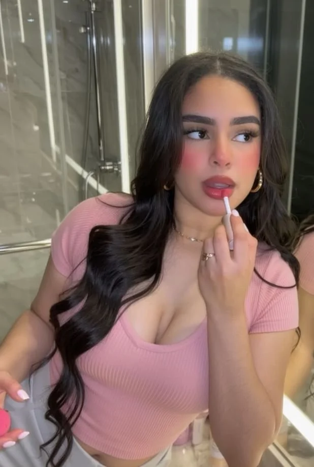 Young woman with long dark hair, wearing a pink top, applying lipstick in front of a mirror in a bathroom or dressing area.