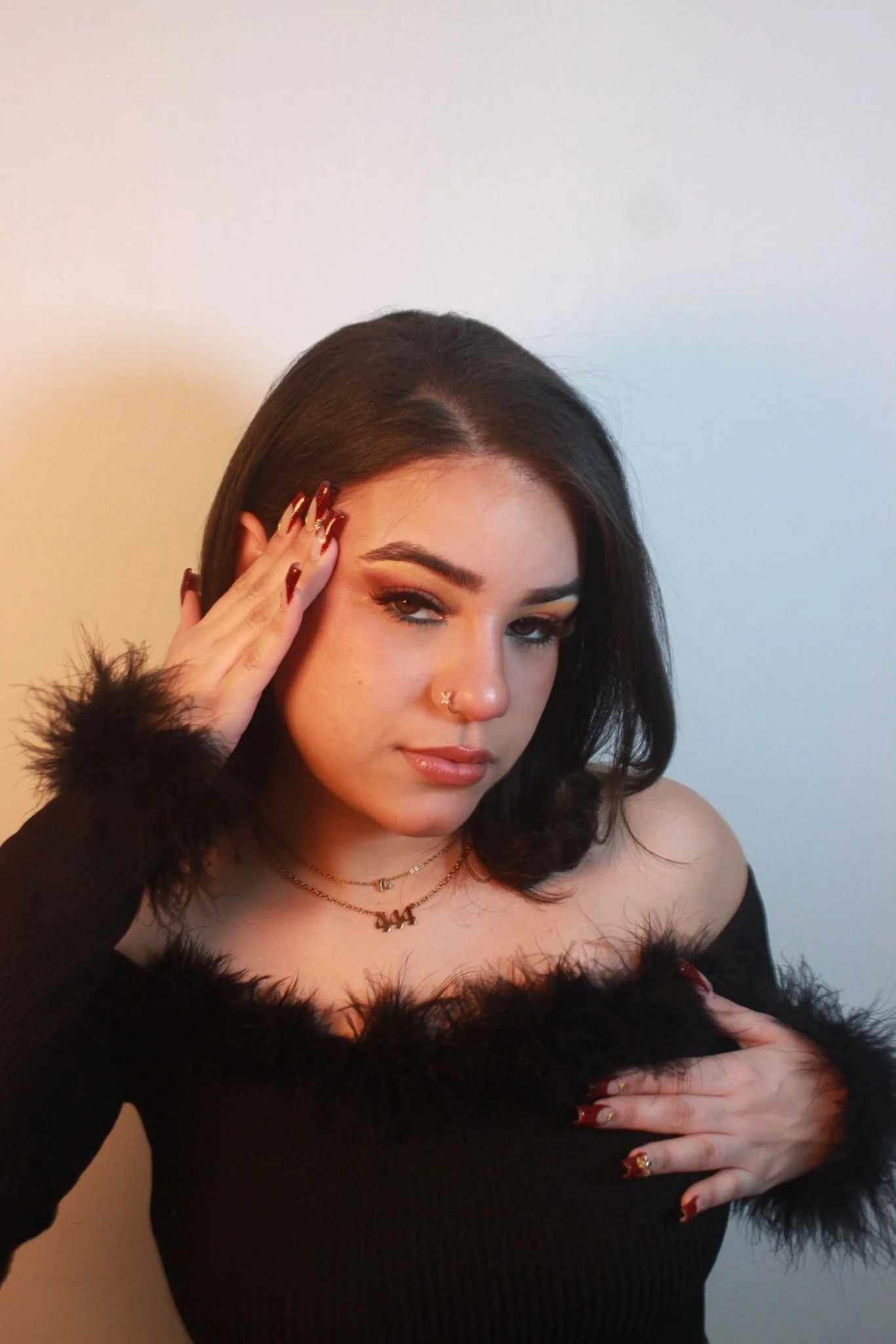 A woman with dark hair and makeup wearing a black off-the-shoulder top with fur trim, jewelry, and red nail polish posing against a neutral background.