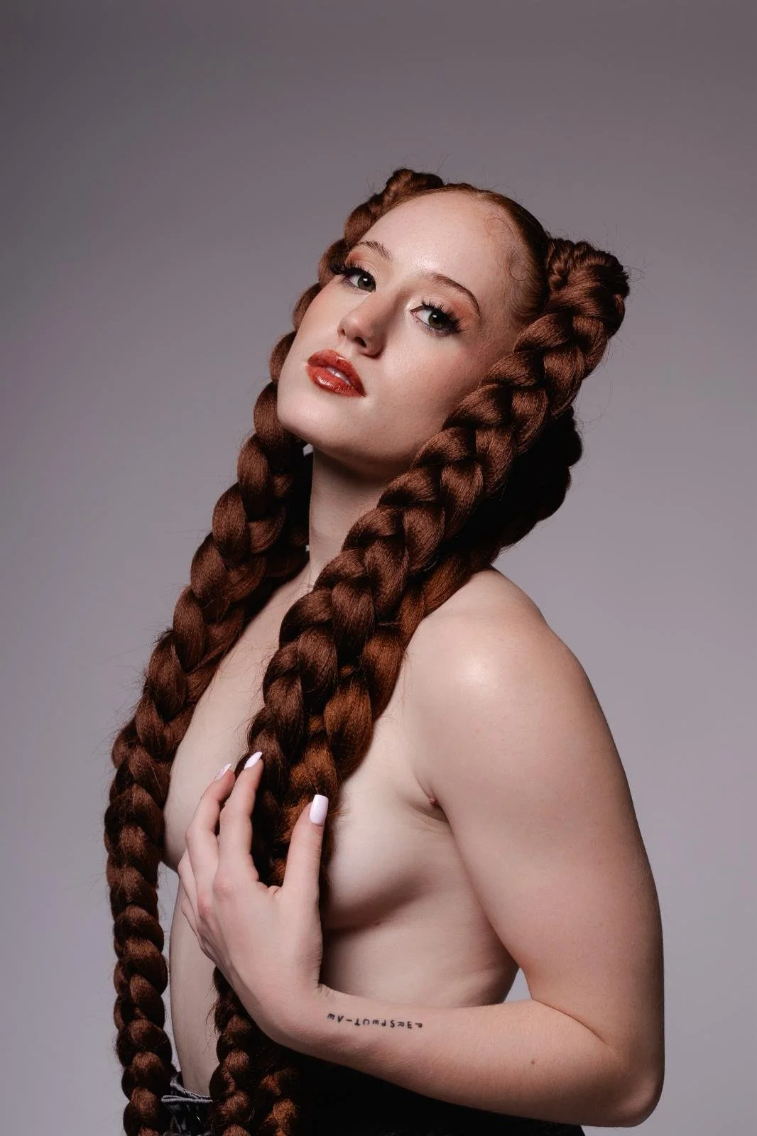 A woman with long, thick, braided red hair, holding one braid with her hand, looking at the camera with makeup and glossy lips, against a plain gray background.