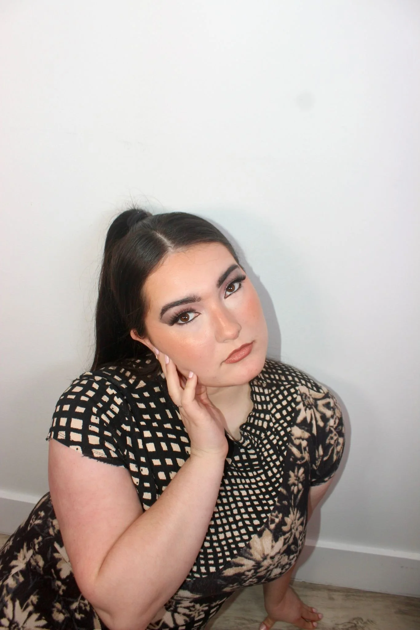 Portrait of a woman with dark hair in a high ponytail, wearing a black and beige patterned dress, posing against a plain white wall.