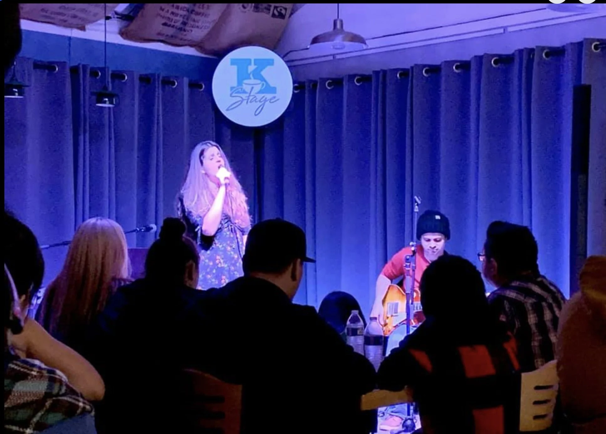 A singer with long hair holding a microphone on stage, performing in front of an audience seated at tables, with a guitarist sitting nearby. The stage has dark curtains, and a round sign with the text 'K Staje' hangs above.
