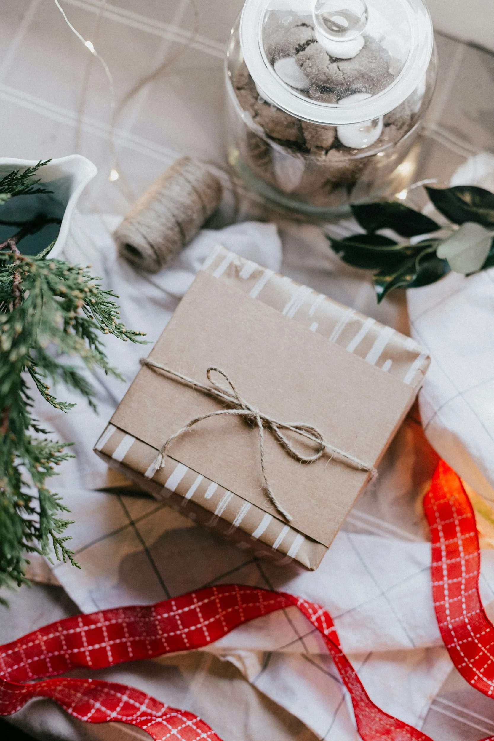 A wrapped gift box with brown paper and white striped paper, tied with a twine bow, surrounded by holiday decorations including a pine branch, red ribbon, a jar of cookies, and a spool of twine.