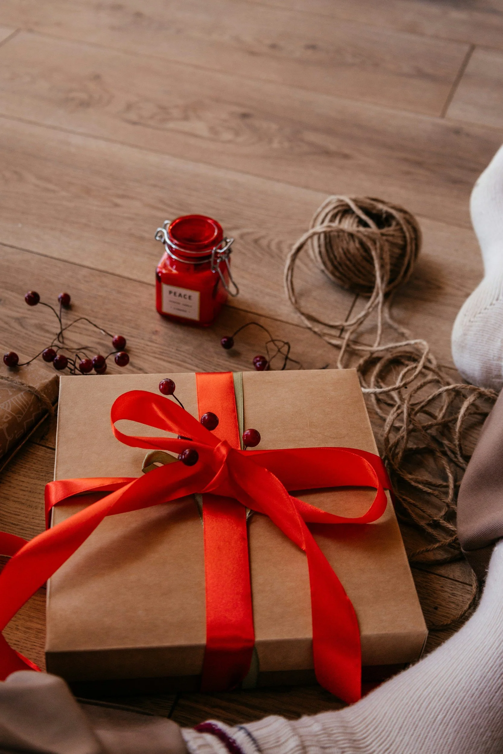 A wrapped gift box with a red ribbon on a wooden floor surrounded by Christmas decorations including a string of red berries, a small jar labeled "Peace," and some twine.