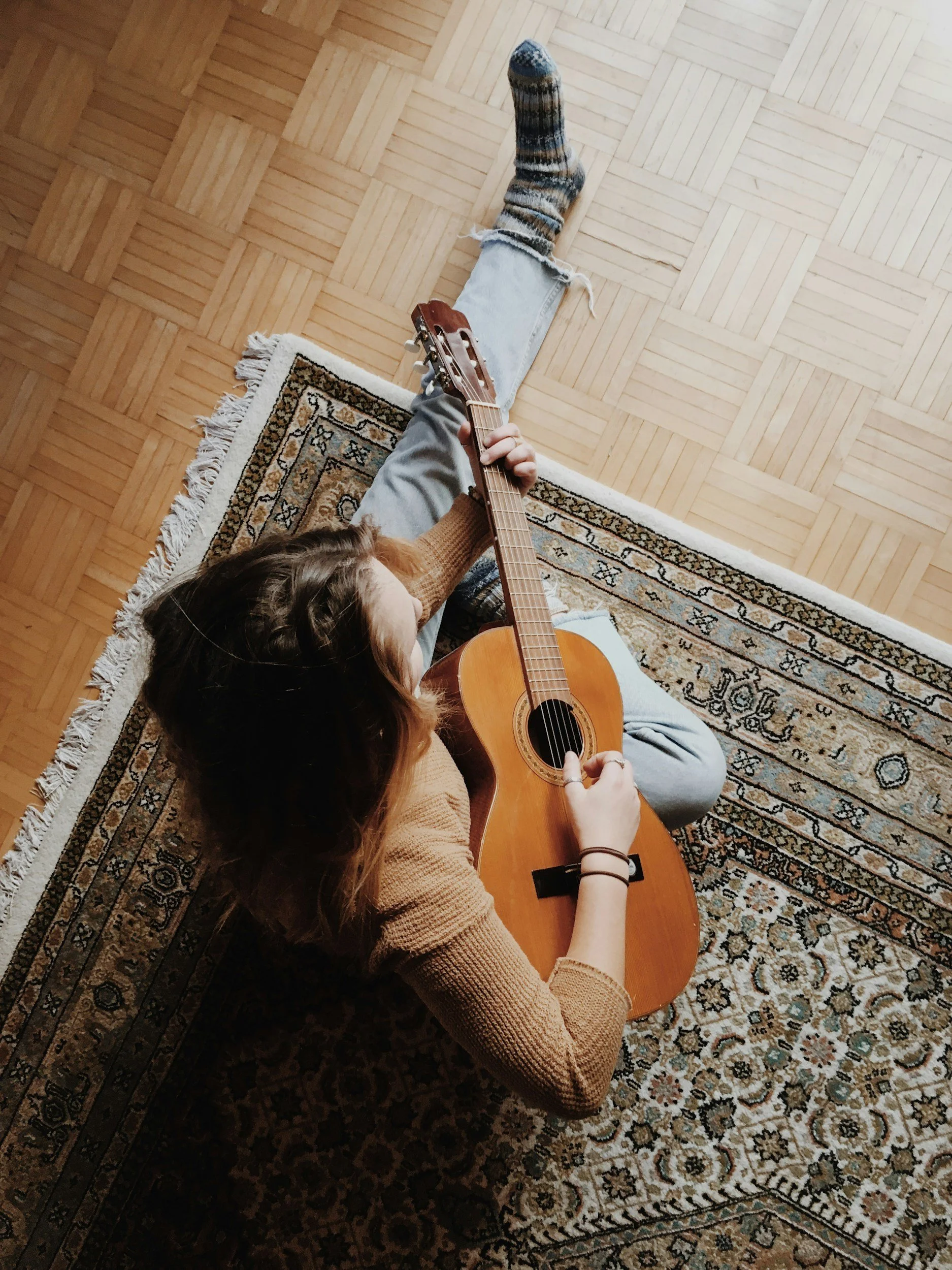 A person sitting on a patterned rug on a wooden floor, playing an acoustic guitar.