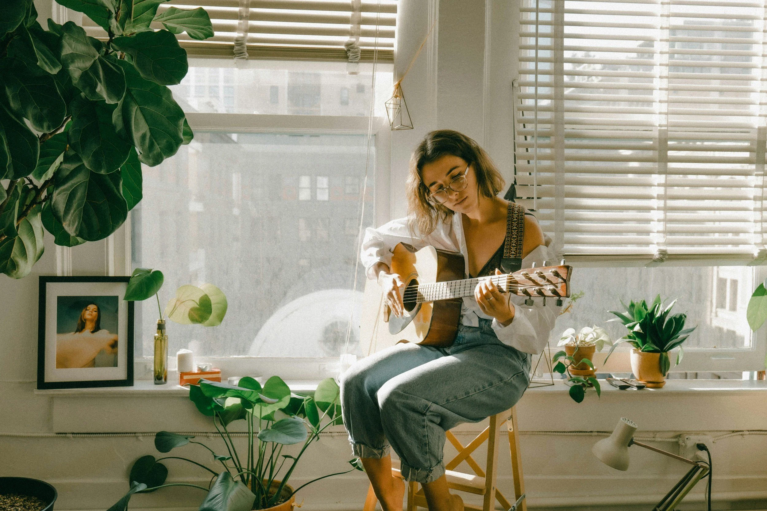 Woman sitting on a stool playing an acoustic guitar in a bright room with large windows, surrounded by houseplants and framed photos.