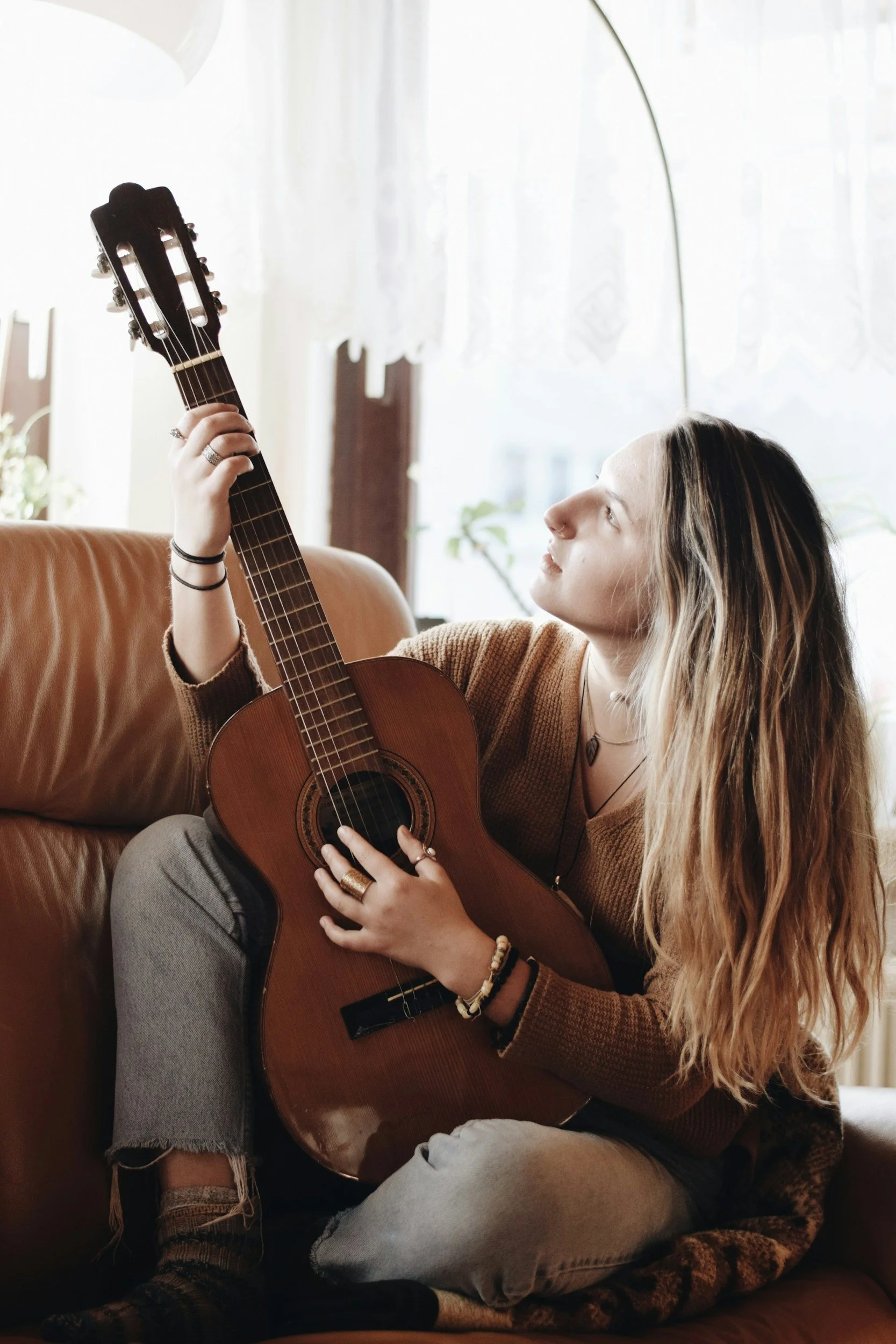 Young woman with long, wavy hair sitting on a leather couch, holding an acoustic guitar, looking thoughtfully upward in a cozy, well-lit room.