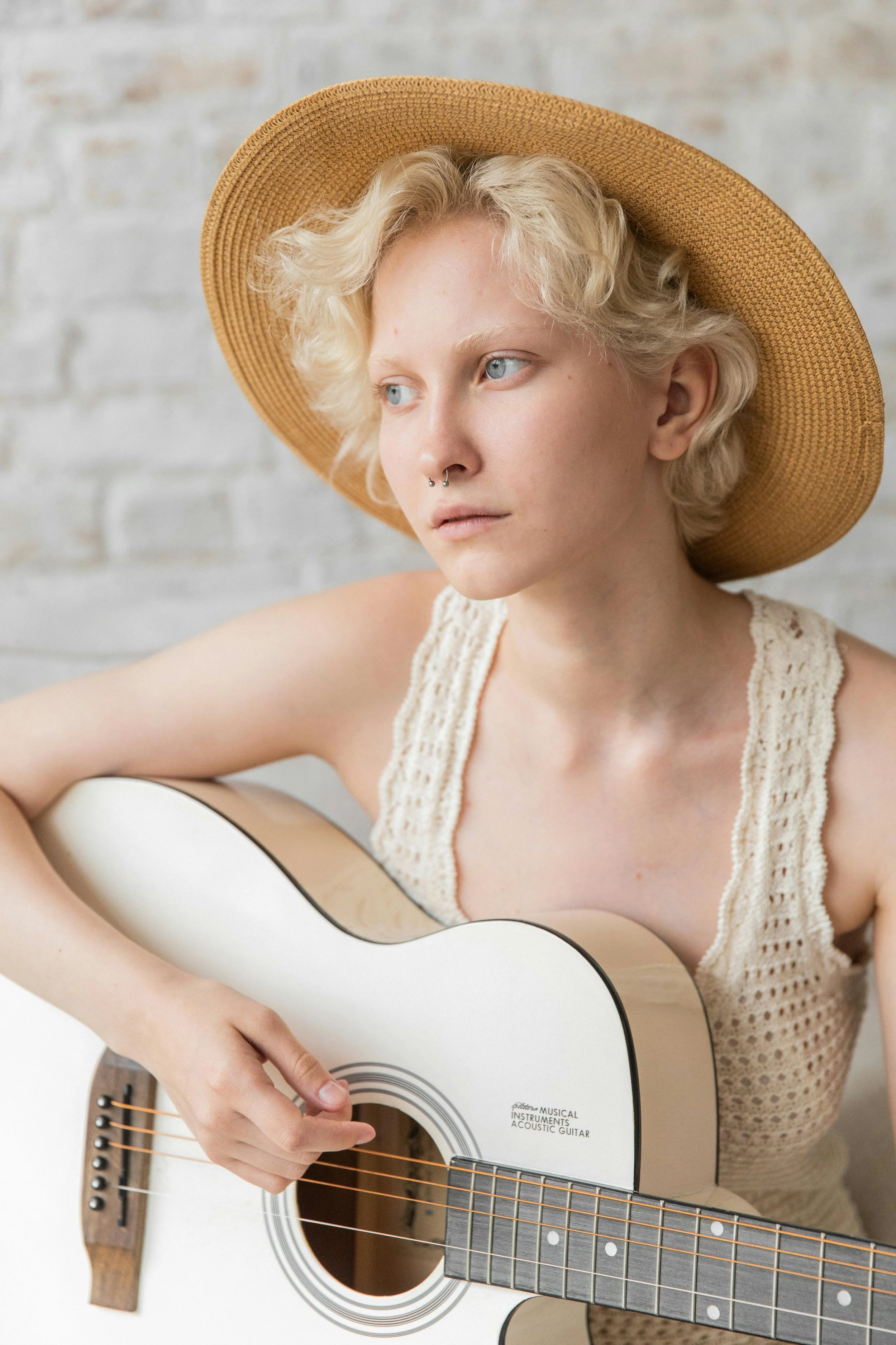 A young woman with short blonde curly hair, wearing a wide-brimmed straw hat and a cream-colored crochet tank top, is playing an acoustic guitar.