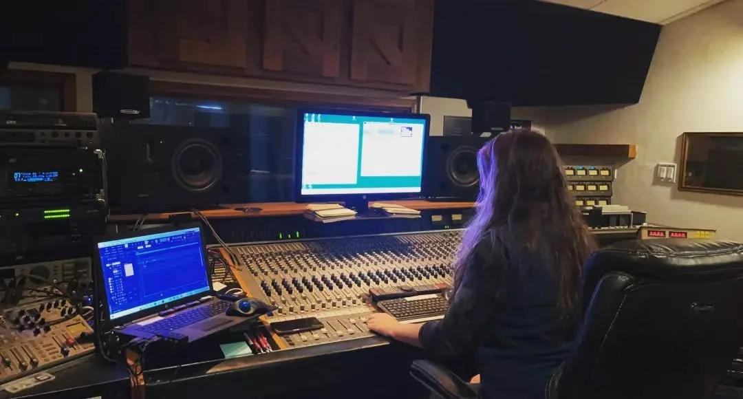 A woman working at a large audio mixing console in a recording studio with various monitors, computers, and audio equipment.