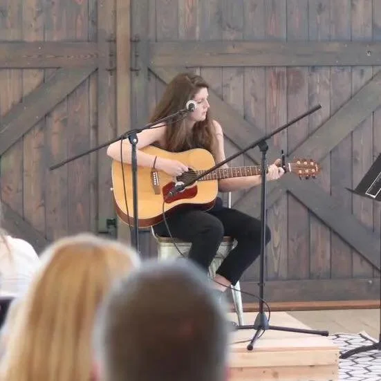 Young woman playing acoustic guitar and singing into a microphone on a stage with a wooden backdrop.