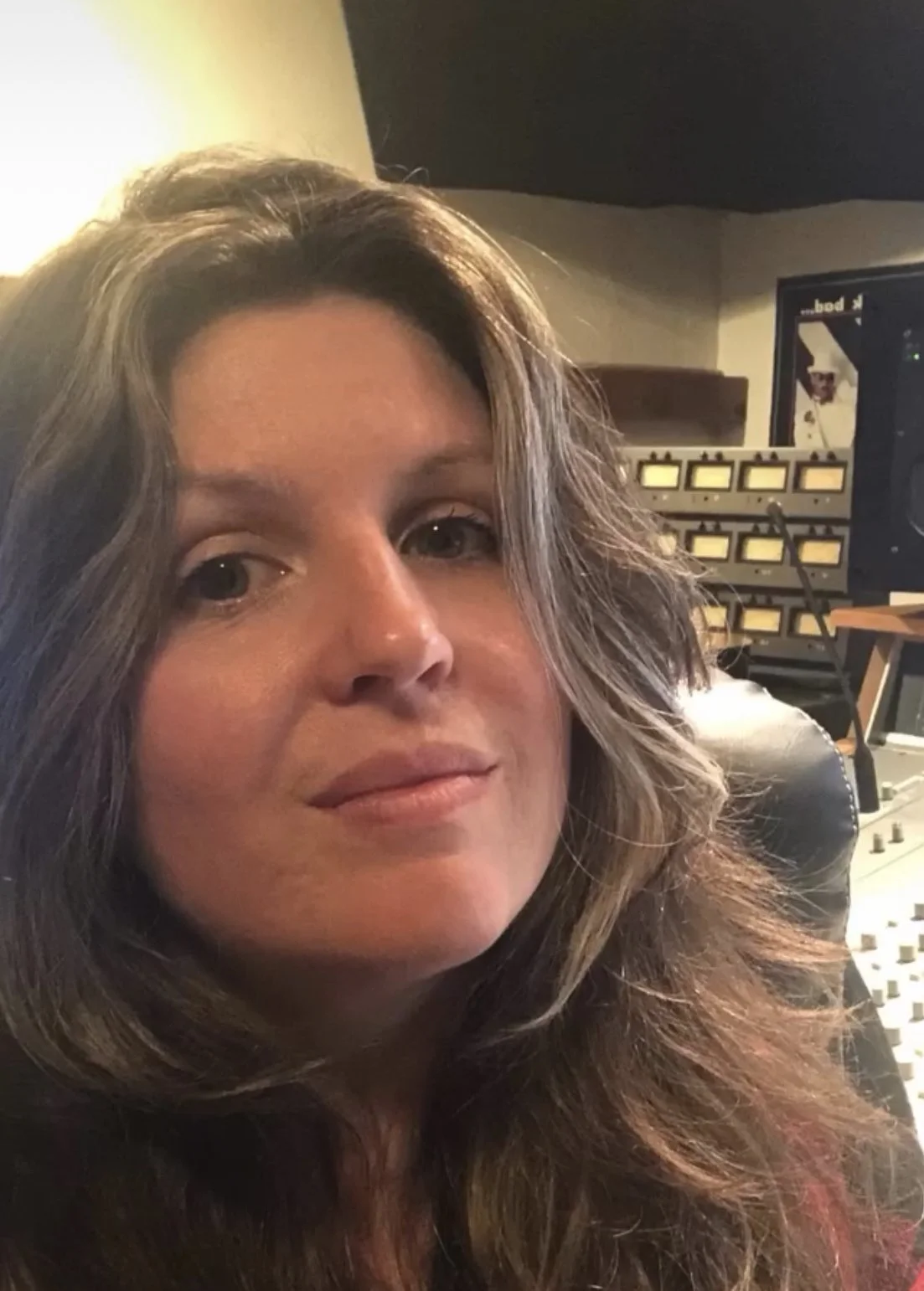 Close-up of a woman with wavy, shoulder-length hair smiling softly, in a recording studio with audio equipment in the background.