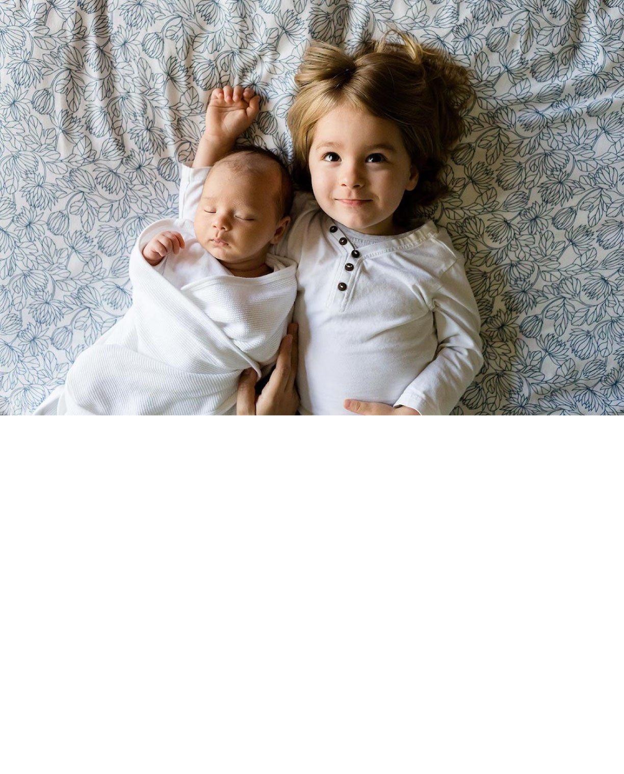 A young girl lying next to a peacefully sleeping baby on a bed with a floral patterned sheet.