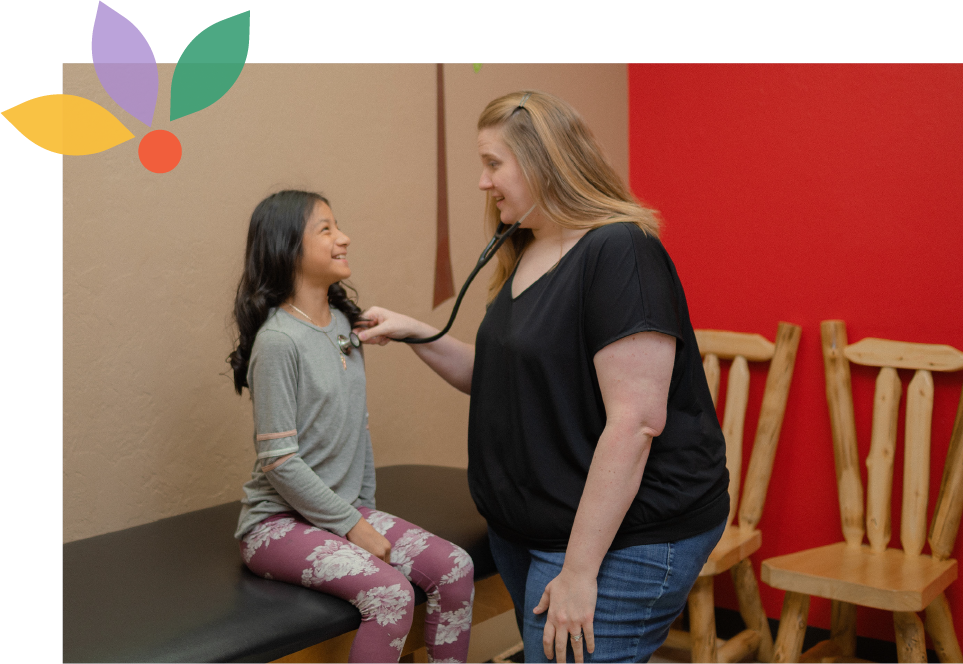 A young girl sitting on an examination table in a doctor's office, smiling at a female healthcare professional who is listening to her chest with a stethoscope.