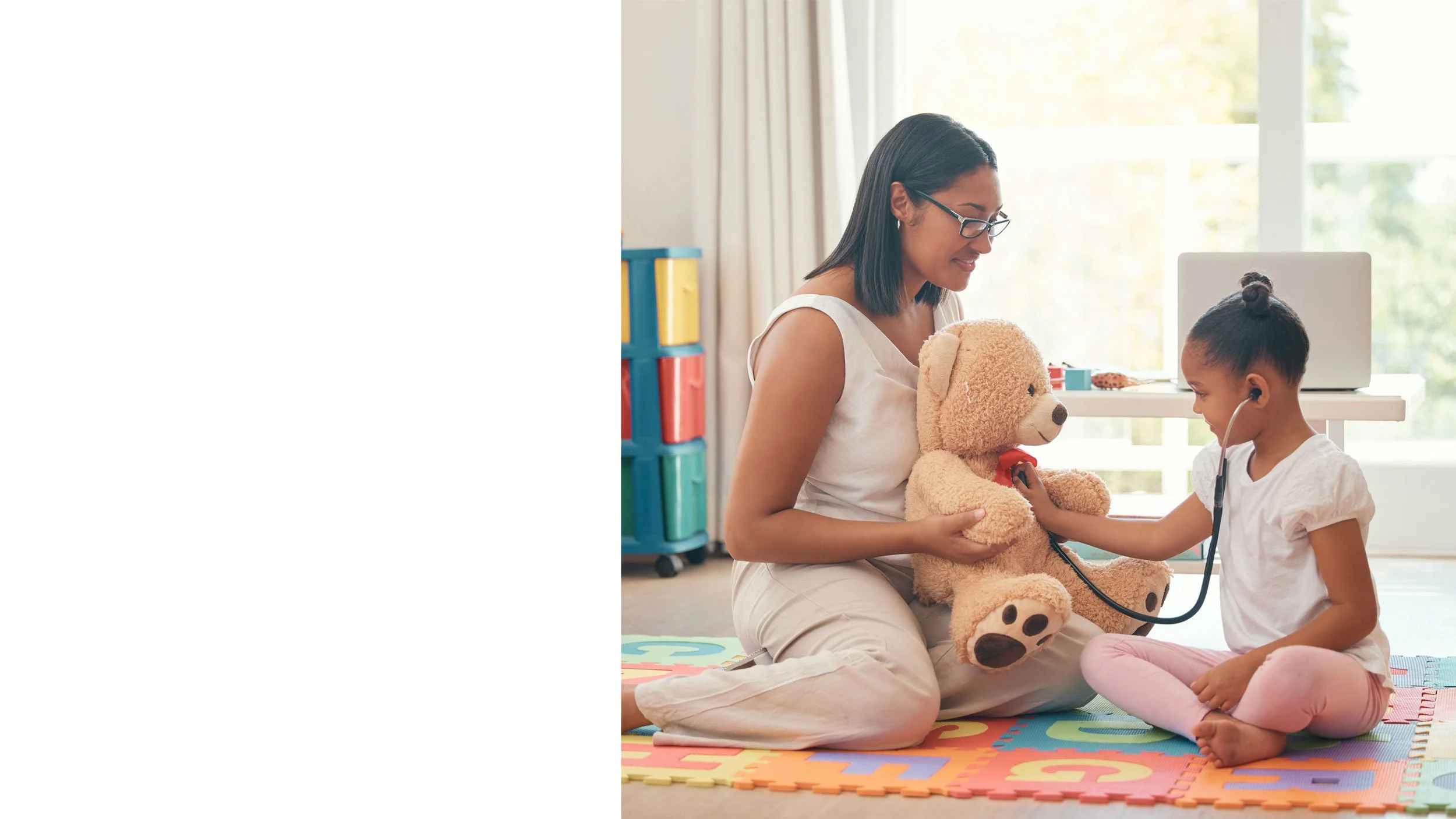A woman smiling down at a young boy who is looking at something off-camera, both wearing white tops, in a close-up indoor setting.