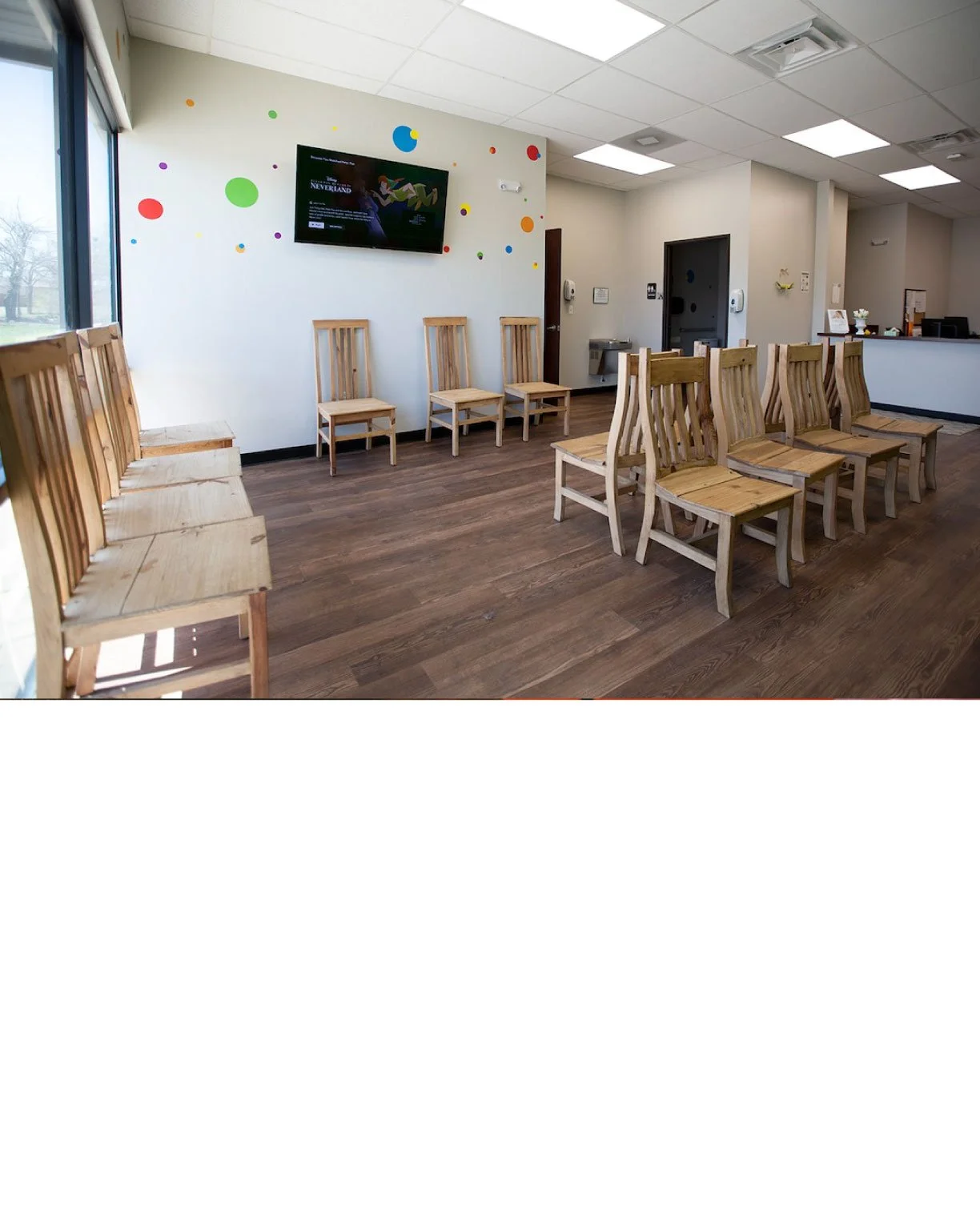 Waiting area with wooden chairs arranged in rows inside a room, a large window on the left, a TV on the wall, and a reception desk in the background.