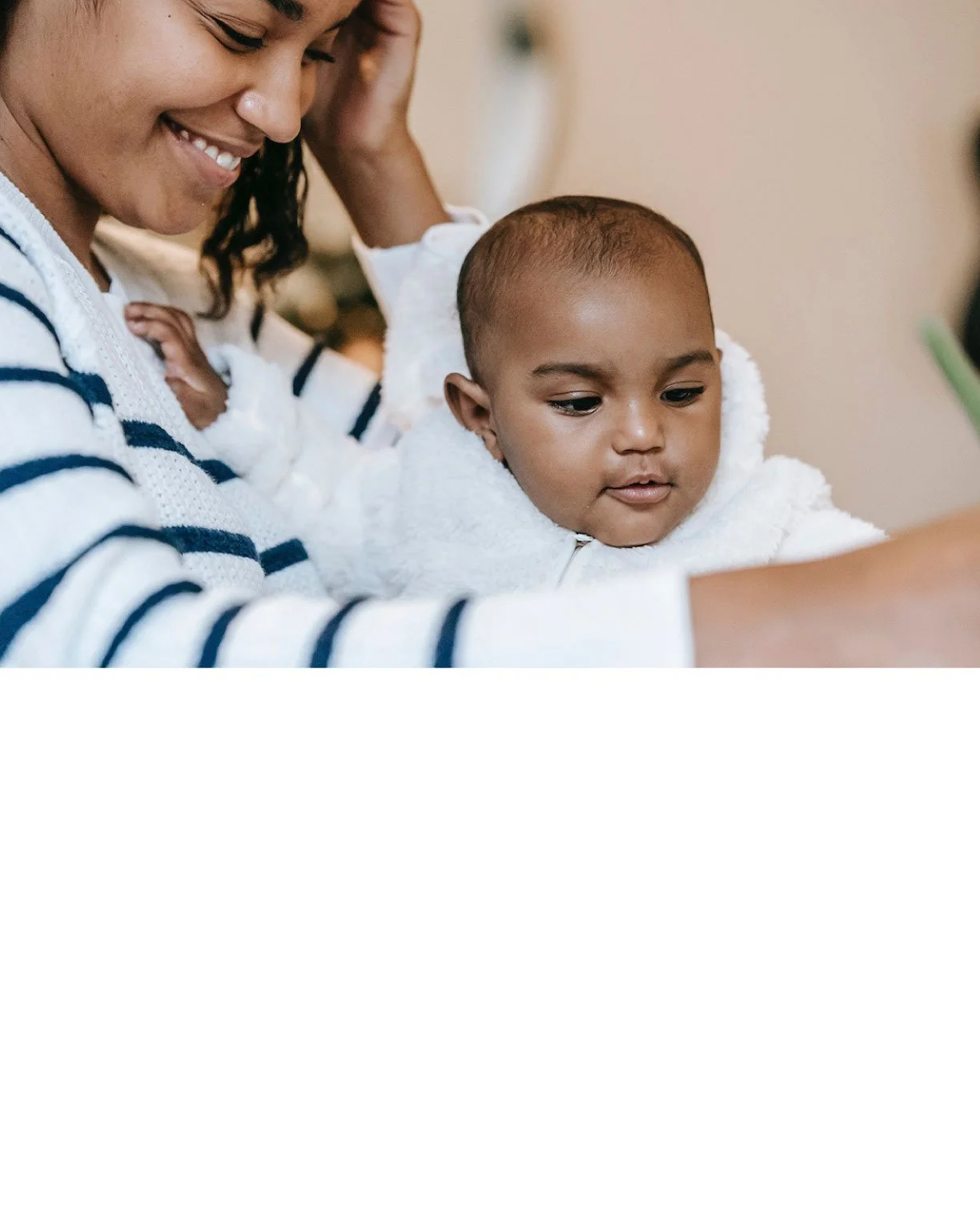 A woman with dark hair and a striped sweater smiling while holding a young child with short hair and a white hoodie, both looking at something in front of them.