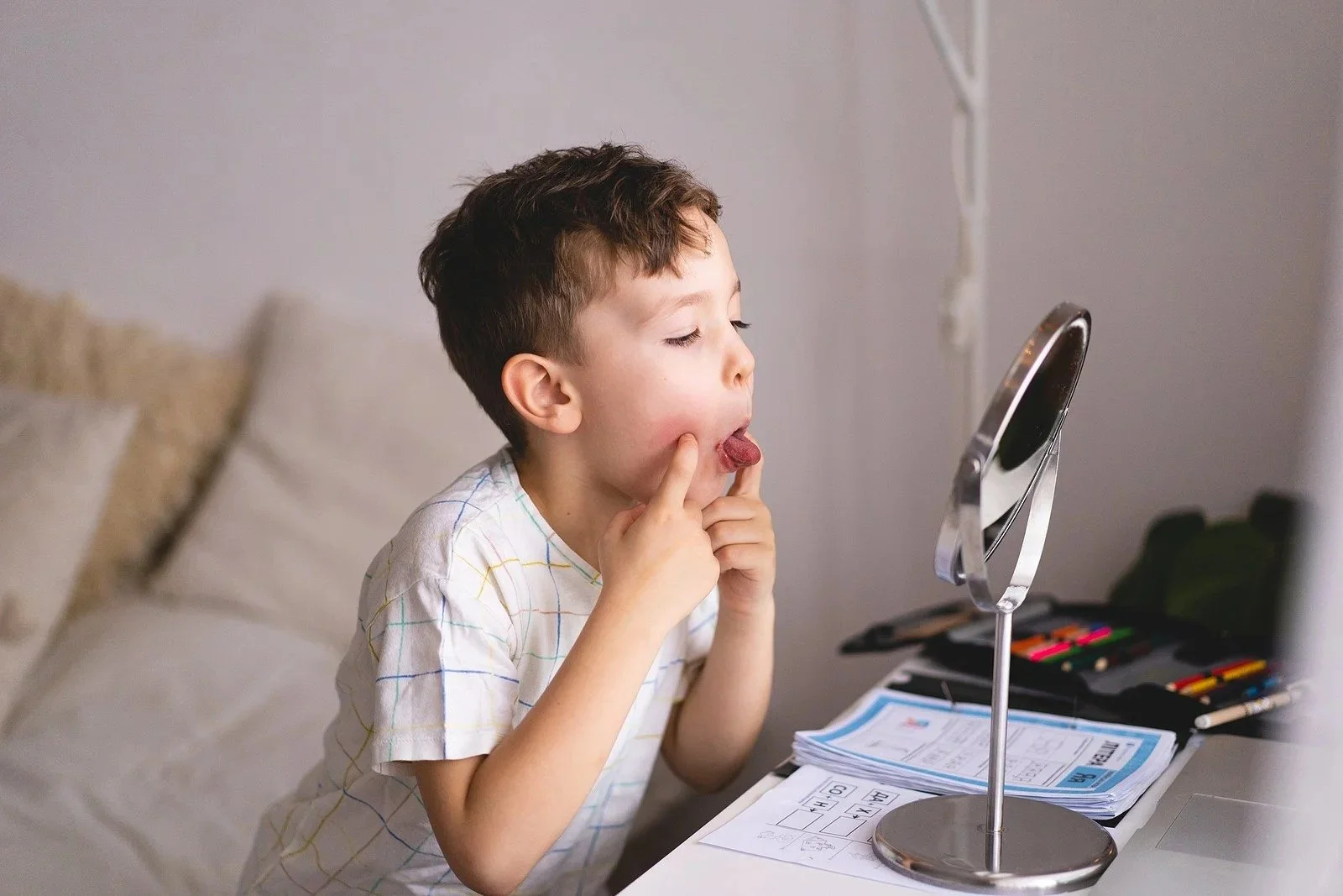 A young boy looks at himself in a mirror with his tongue out and fingers on his cheeks, sitting at a desk with papers and art supplies.