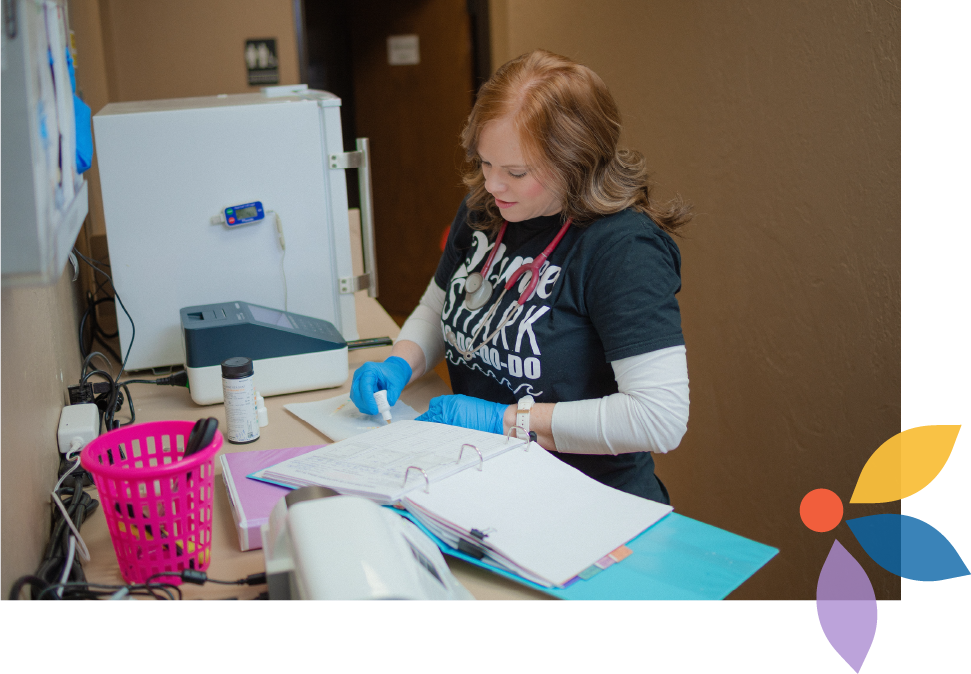 A female healthcare worker wearing blue gloves and a stethoscope around her neck filling out paperwork at a desk in a medical office.