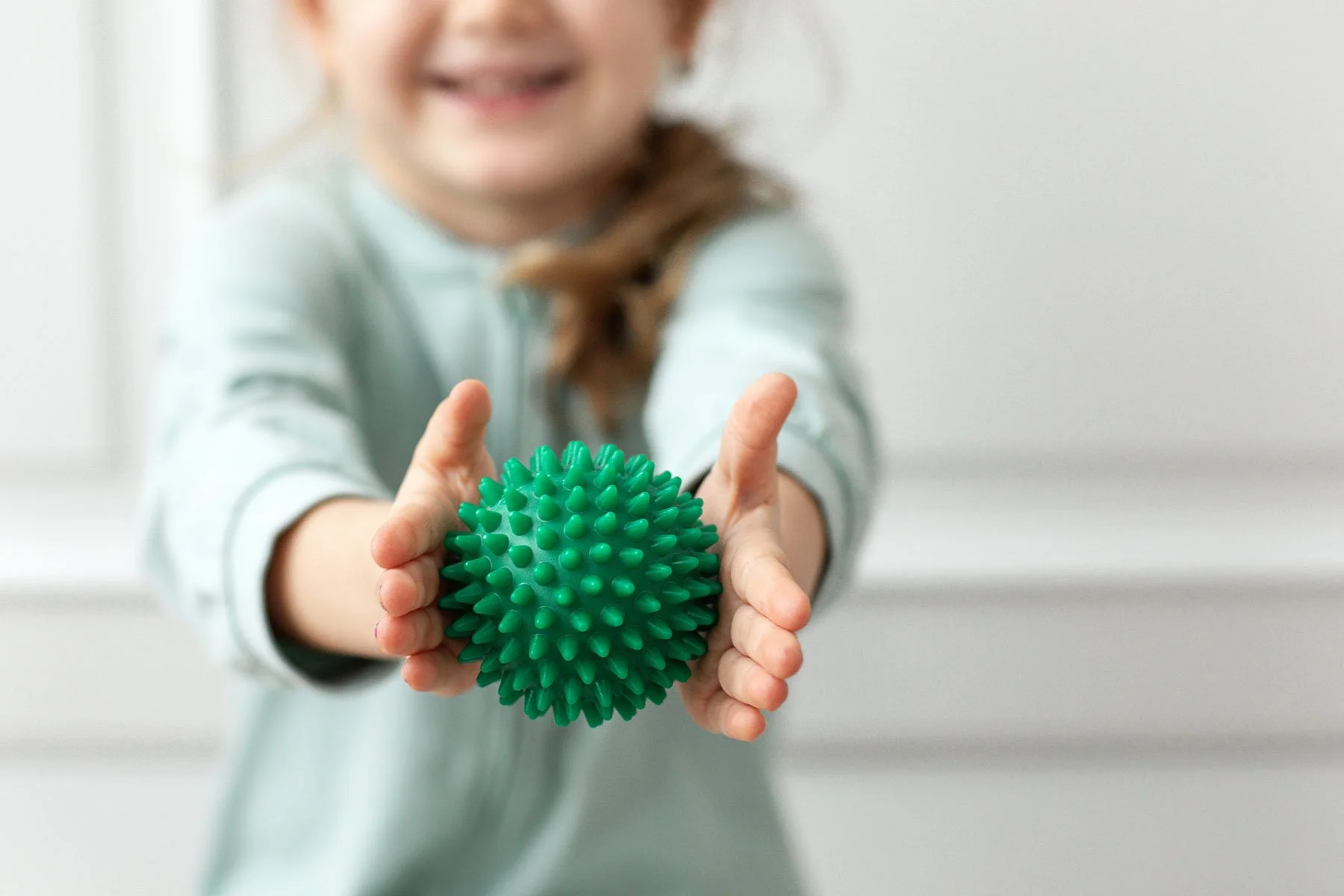 A smiling child holding a green spiky massage ball towards the camera.