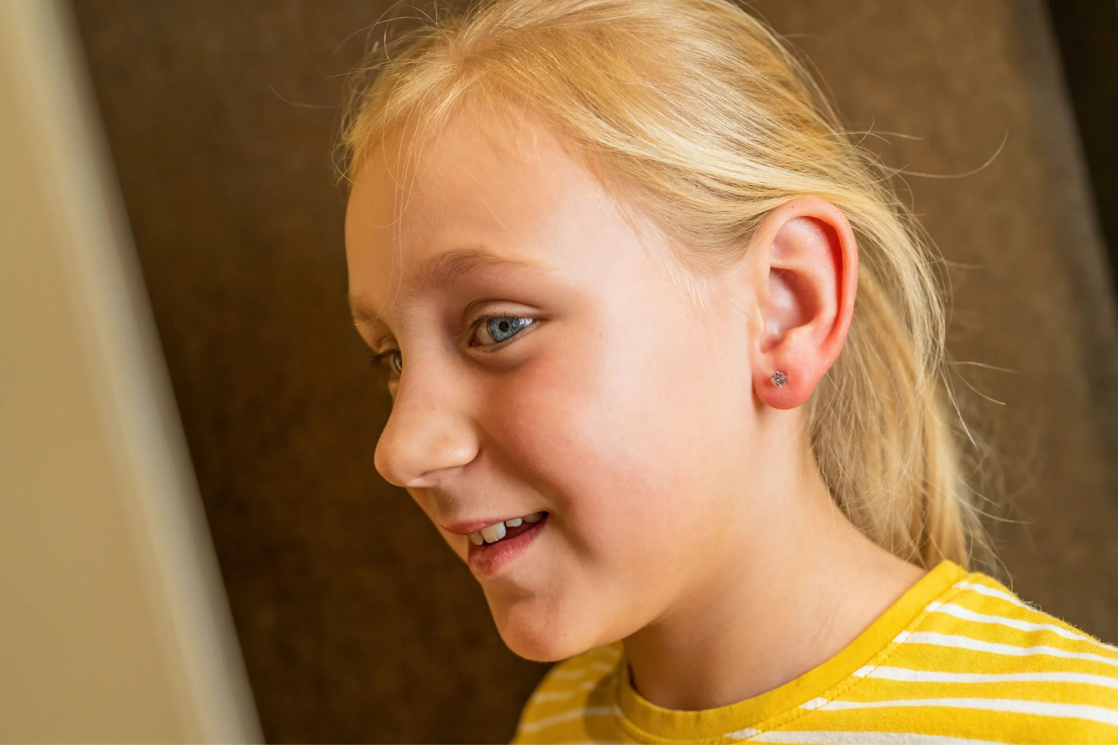 A close-up of a young girl with blonde hair, blue eyes, wearing a yellow and white striped shirt, smiling and looking at a screen.