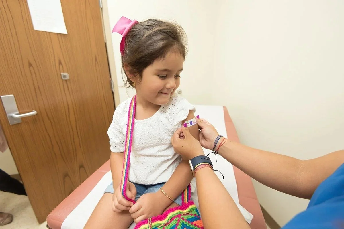 A young girl sitting on an examination table, getting a vaccination shot in her upper arm from a healthcare professional.