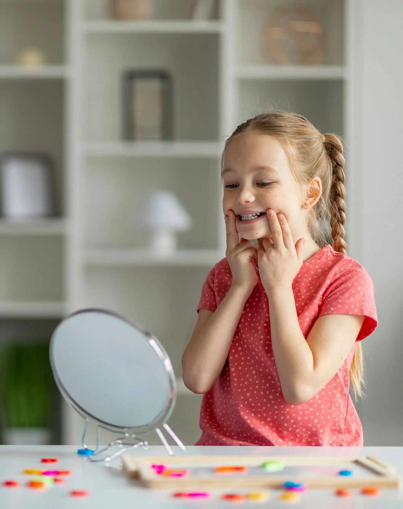 A young girl smiling in front of a mirror at a table with colorful craft supplies.