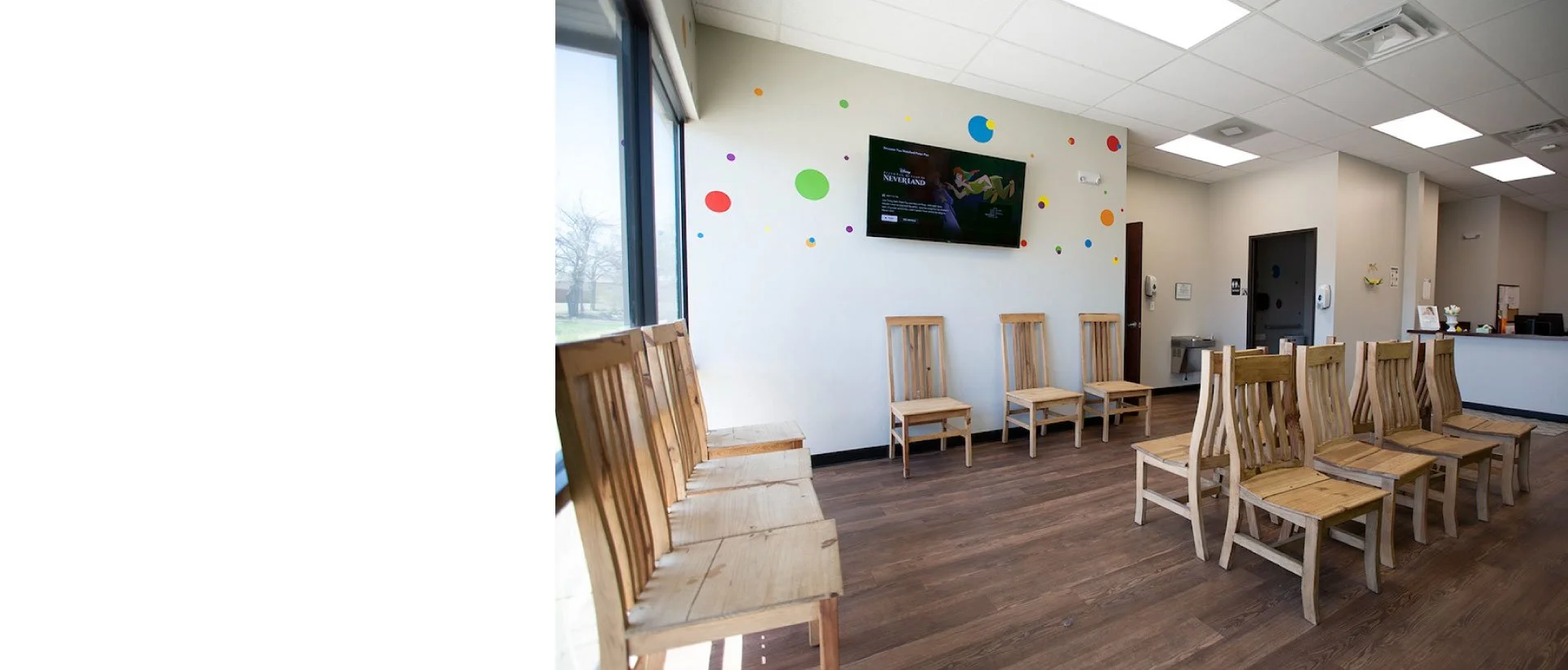 Waiting room with wooden chairs lined up against a wall, colorful dot decorations, and a television screen mounted on the wall.