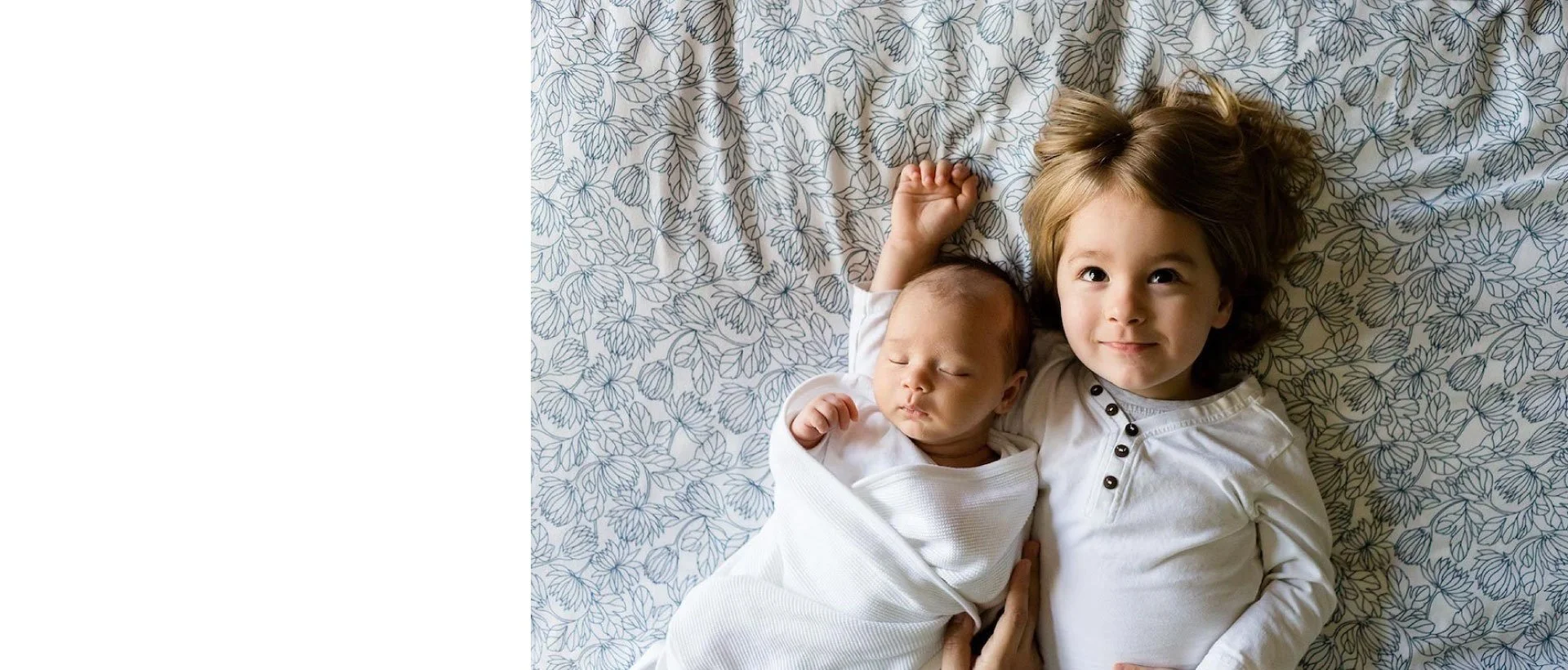 A young girl with light brown, wavy hair lying on a bed next to a sleeping infant wrapped in a white blanket. The girl is smiling slightly and wearing a white shirt with black buttons.