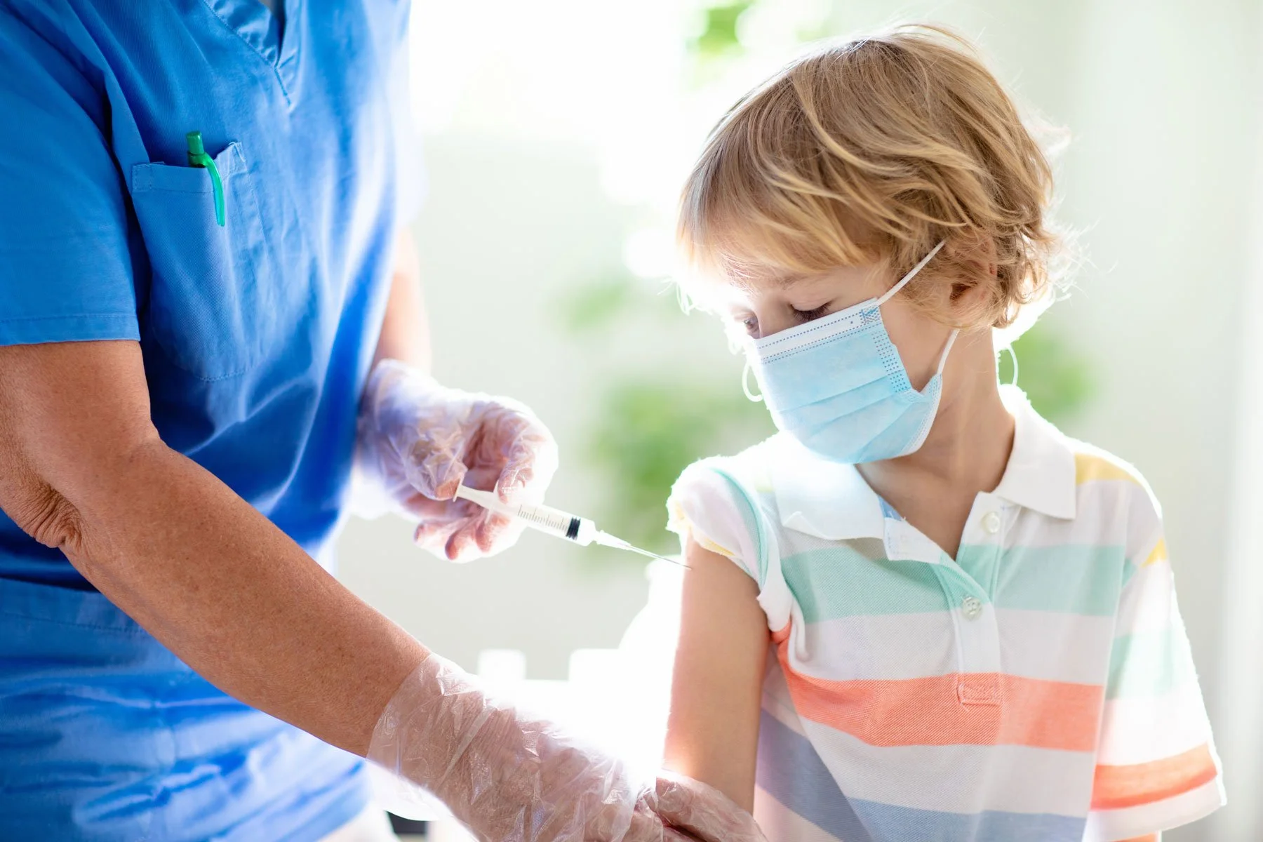 A young boy receiving a vaccination shot from a healthcare professional in a medical setting. Both are wearing face masks, and the healthcare worker is wearing gloves and a blue uniform.