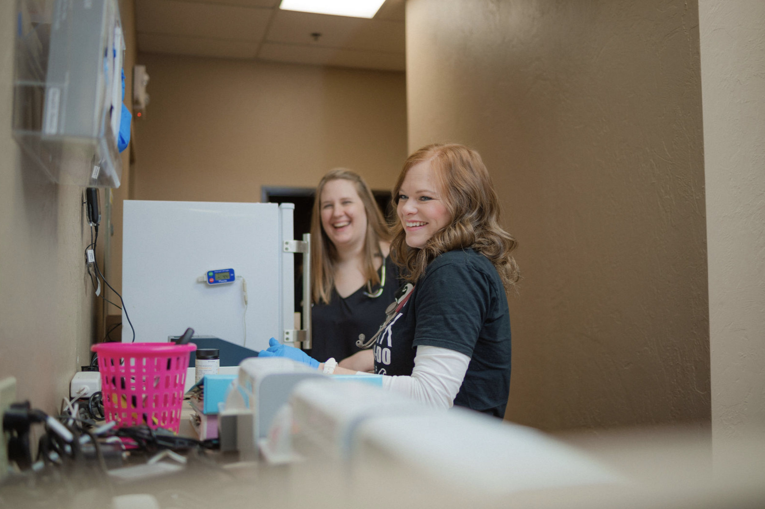 Two friendly pediatric healthcare professionals smile while working in a clinical setting at Just Kids Pediatrics, showcasing a warm and welcoming environment for child-focused medical care.
