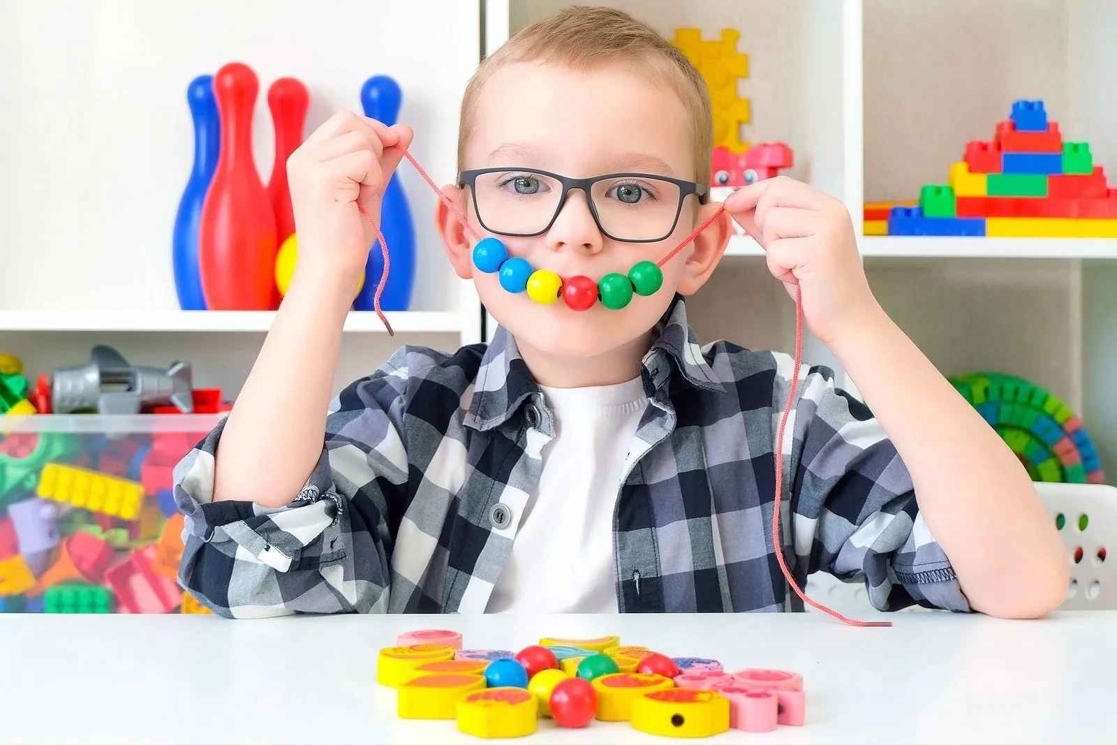 A young boy wearing glasses and a plaid shirt creates a smile with colorful beads on a string, sitting at a table with various toys and game pieces in the background.