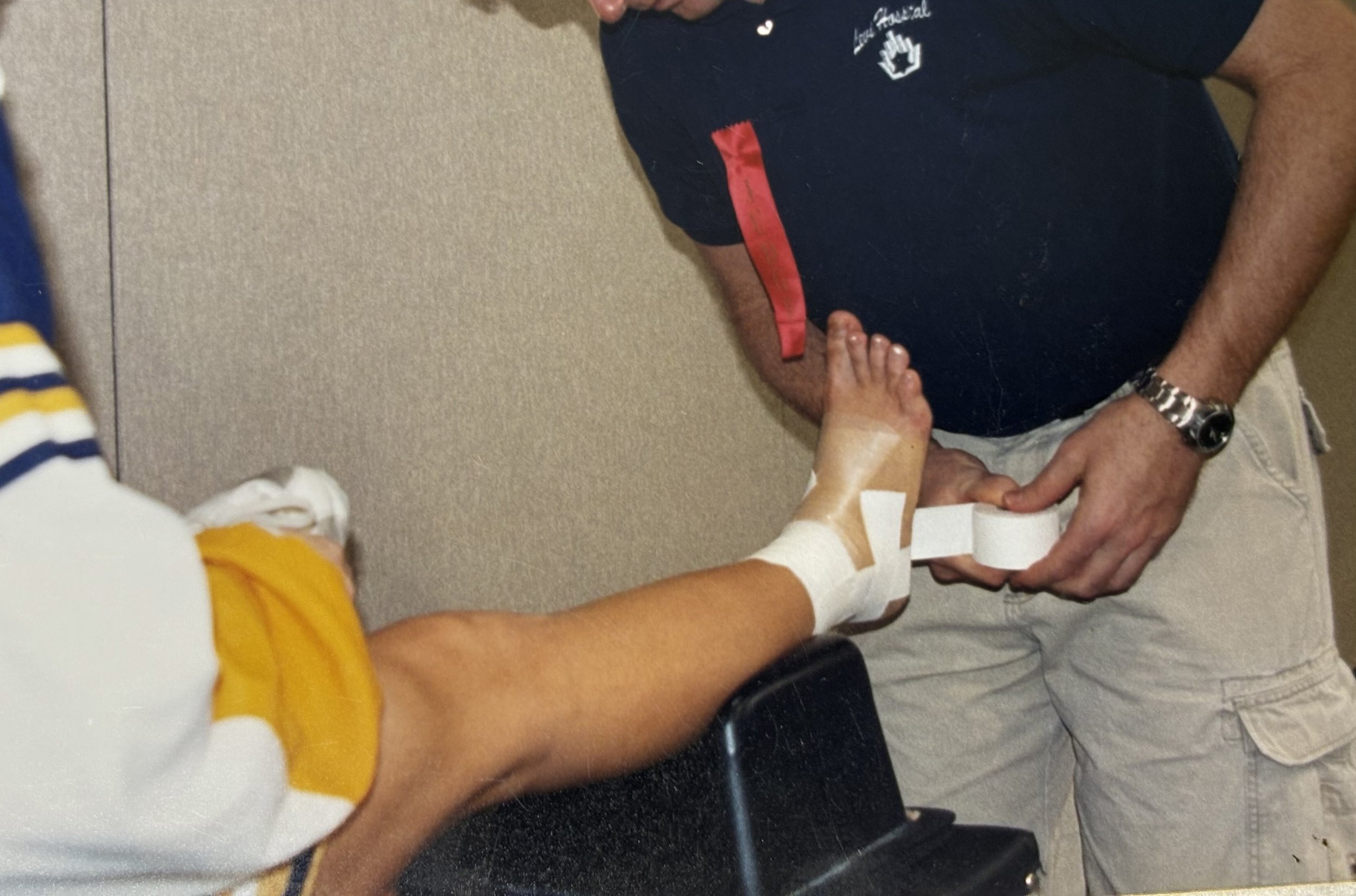 A person is wrapping a bandage around a patient's ankle, who is sitting on a chair, in a medical setting.