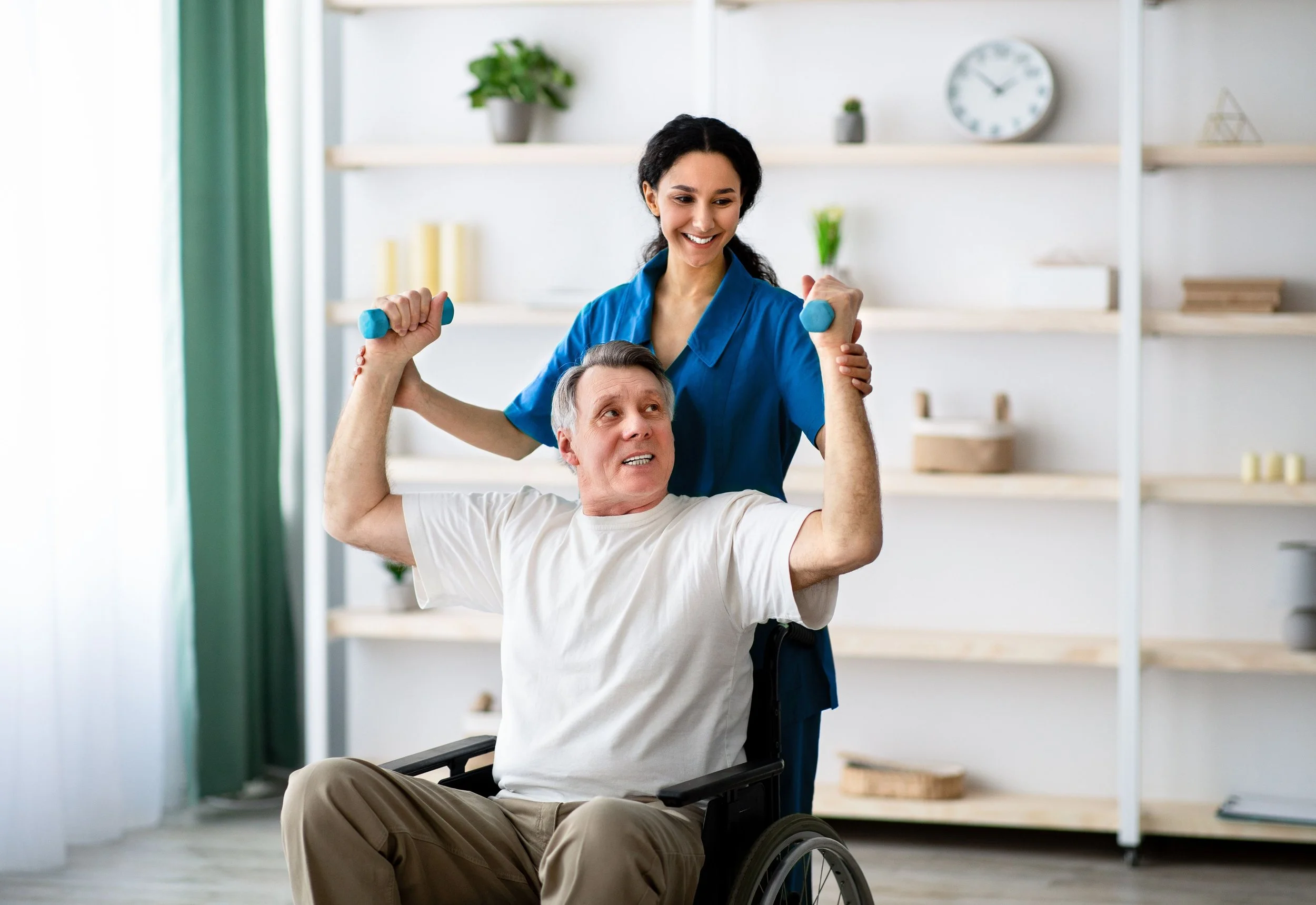 A woman in a blue uniform assisting an elderly man in a wheelchair with arm exercises using small dumbbells.