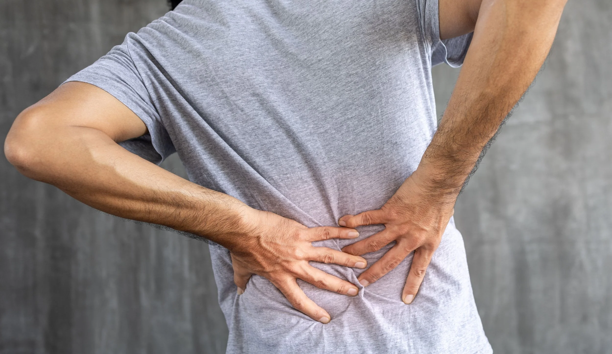 Person wearing a gray t-shirt holding their lower back with both hands, indicating back pain.