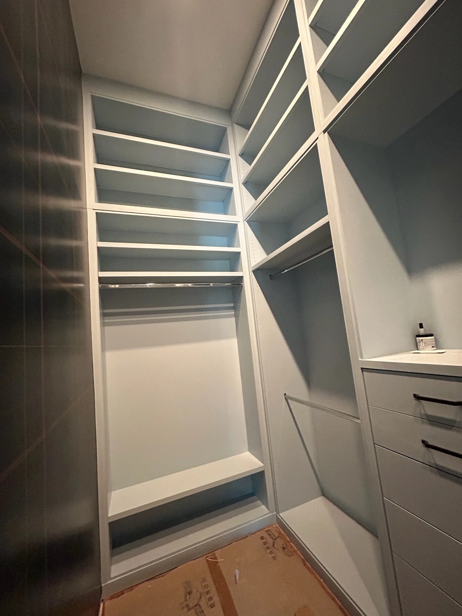 Empty walk-in closet with white shelving and drawers, a hanging rod, and a cardboard box on the floor.