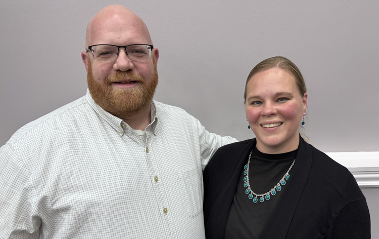 A man with glasses, a bald head, and a red beard, dressed in a white checkered shirt, standing next to a woman with blonde hair, wearing a black blazer and turquoise jewelry, both smiling and posing together against a plain, light gray wall.