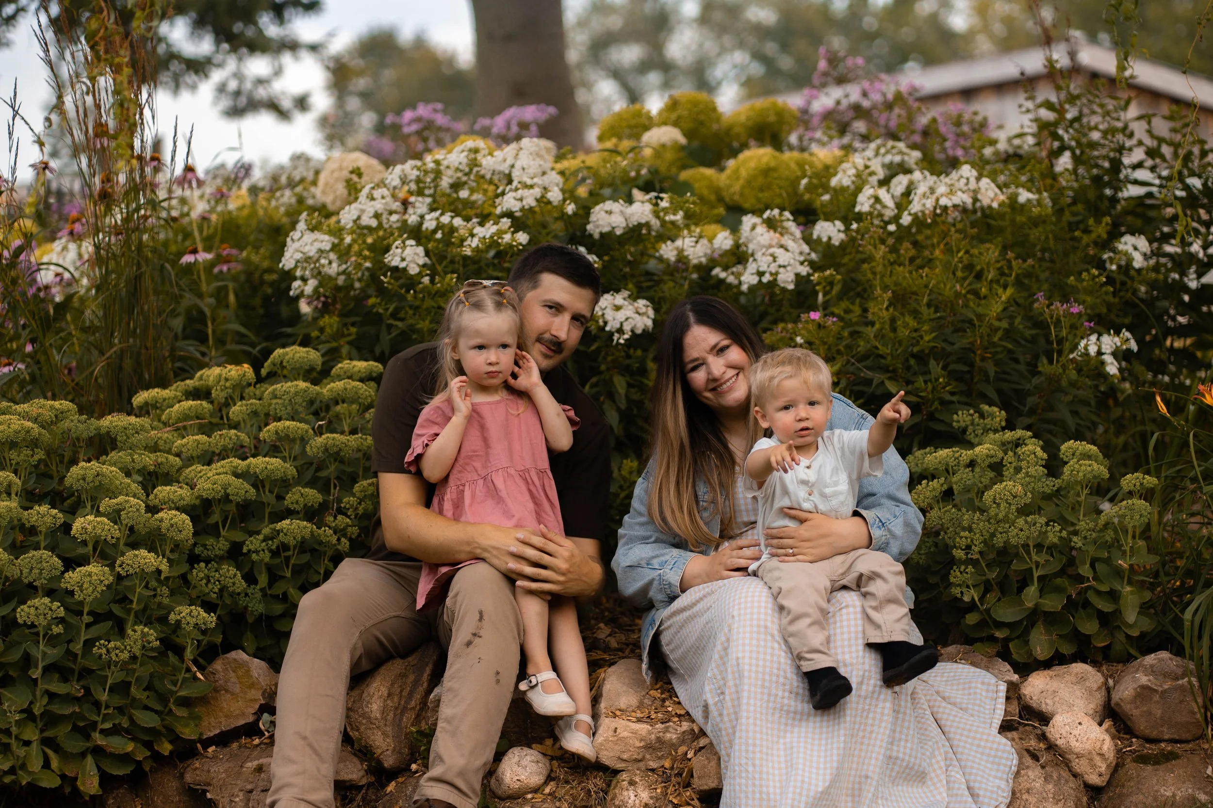 A family of four sitting outdoors among flowers and greenery, with a man, woman, and two young children.