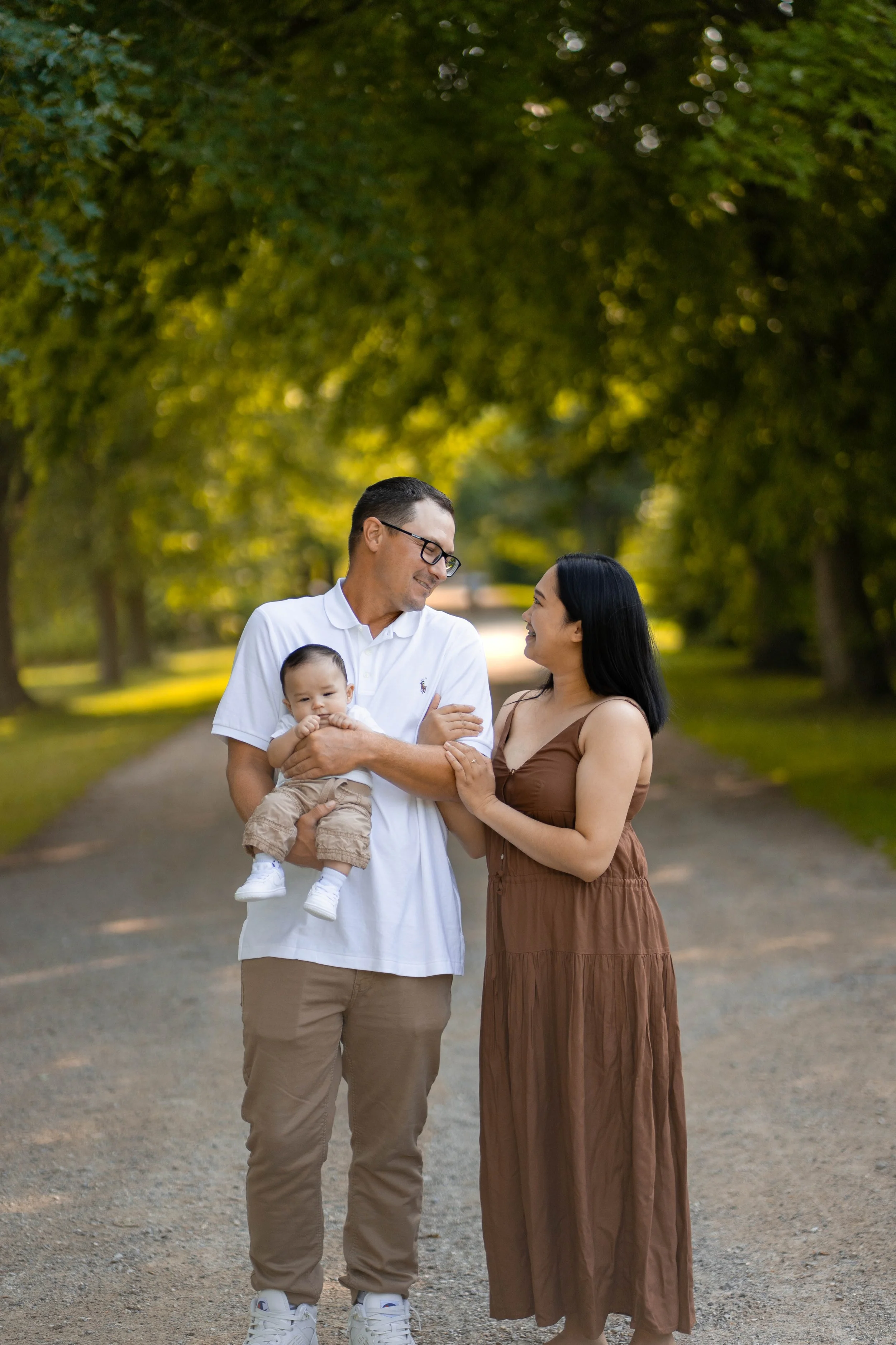 A family of three standing on a dirt path in a park with lush green trees. The man in a white shirt is holding a baby, and the woman in a brown dress is touching his arm. They are smiling and looking at each other.