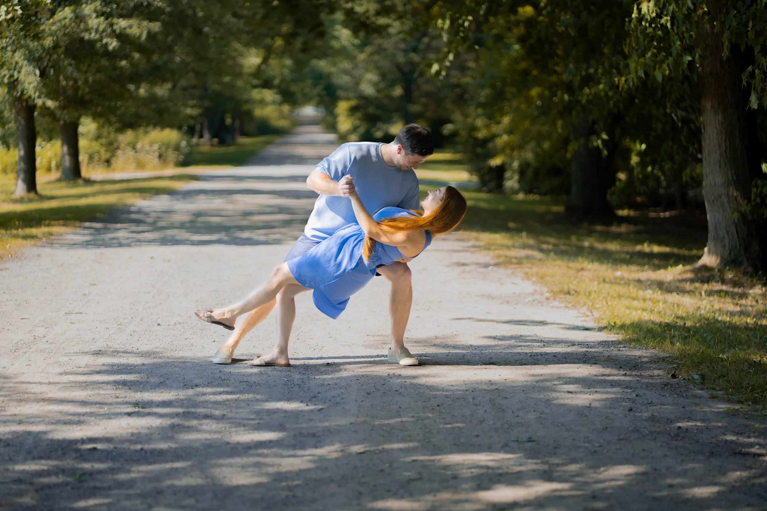 A man in a light blue t-shirt and shorts is holding a woman in a blue dress and sandals as they dance or play on a tree-lined dirt road in a park on a sunny day.
