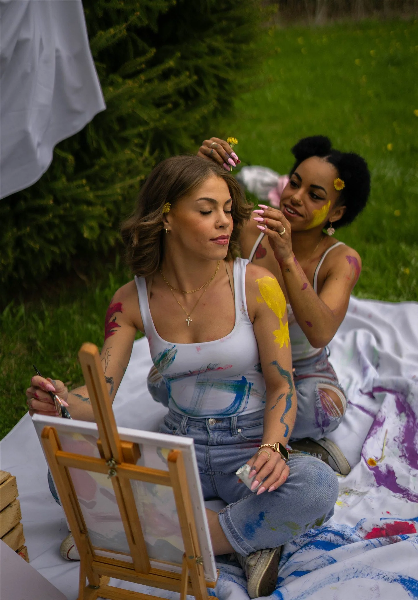 Two women with paint on their bodies sitting on a blanket outdoors, one painting on the other's shoulder while the other closes her eyes, with a small easel, surrounded by greenery.