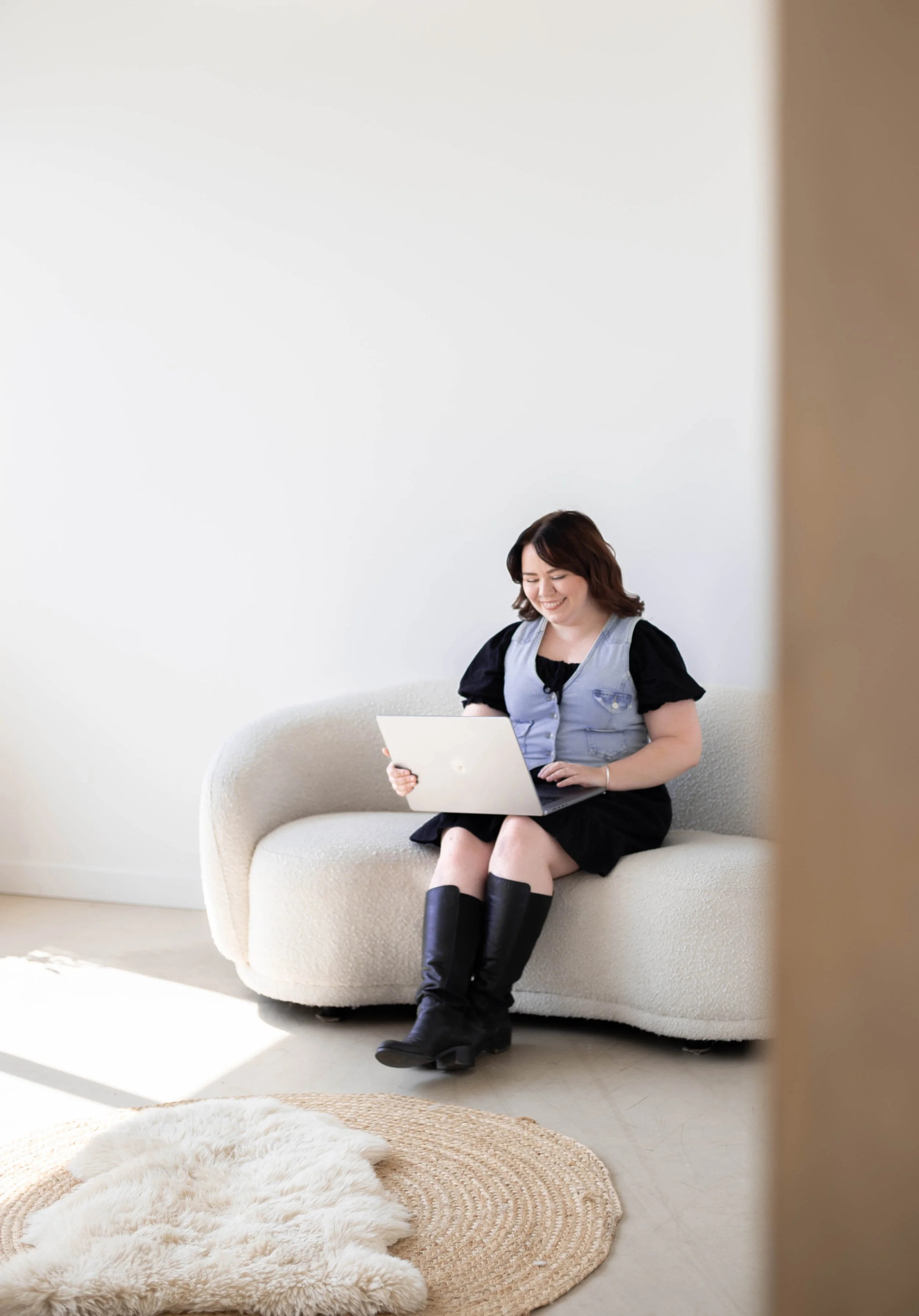 A woman with short brown hair sitting on a cream sofa, smiling and looking at a laptop on her lap in a bright, minimalist room.