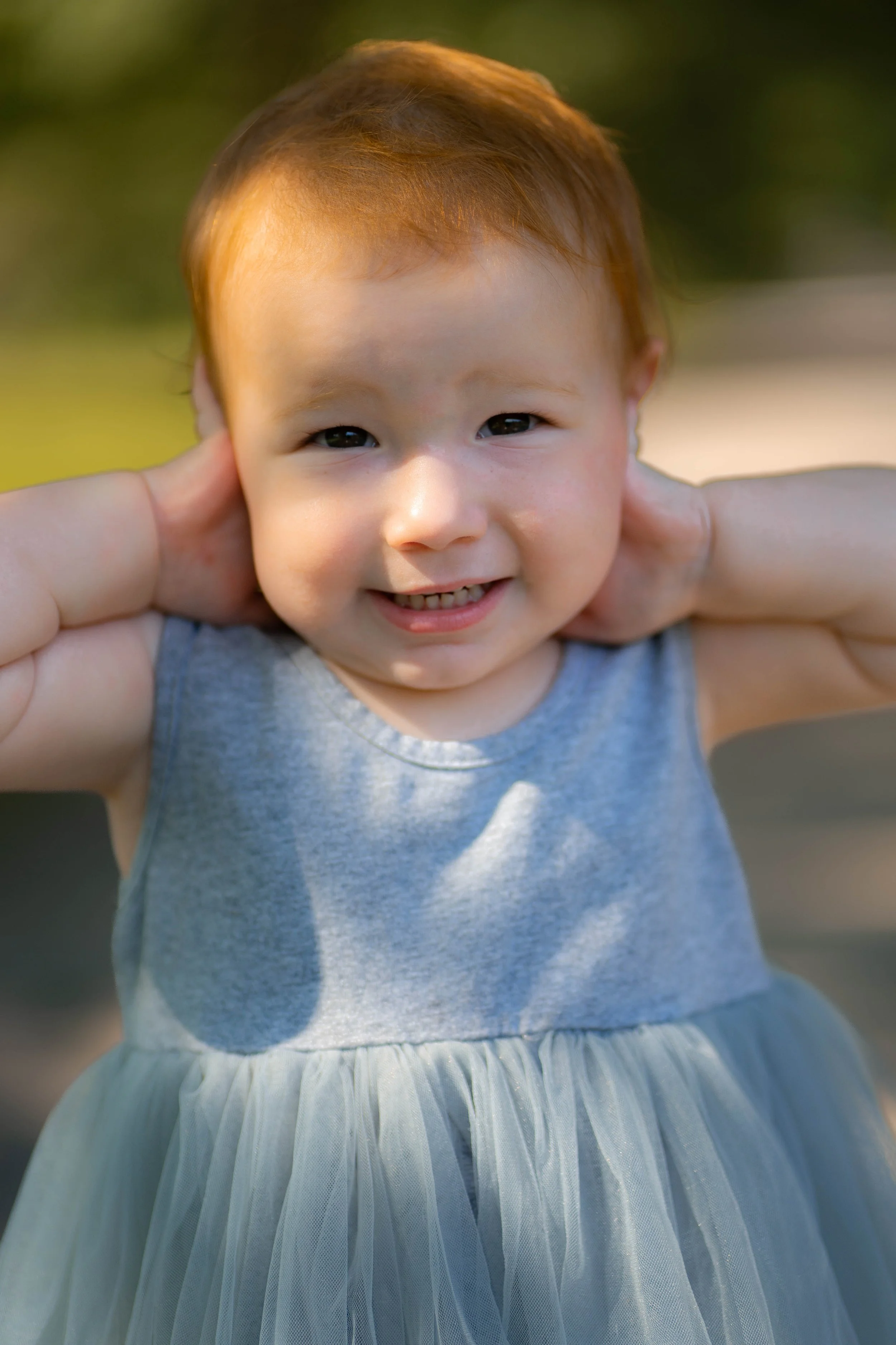 Close-up of a young girl with red hair smiling, wearing a sleeveless gray dress with a tulle skirt, outdoors with blurred greenery in the background.