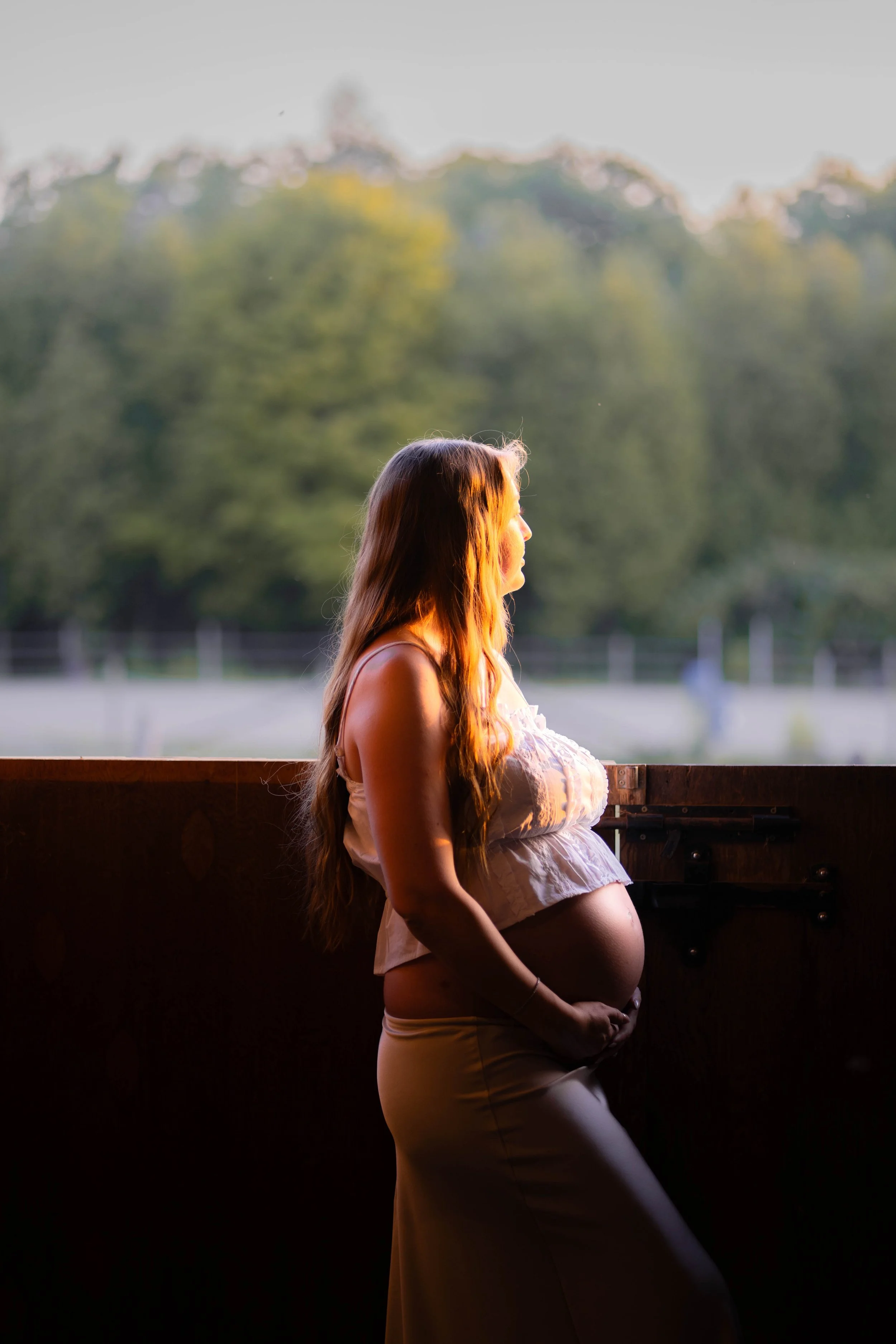 Pregnant woman standing by a wooden railing outdoors, looking to the right with a scenic background of trees and a fence in the distance, during golden hour.