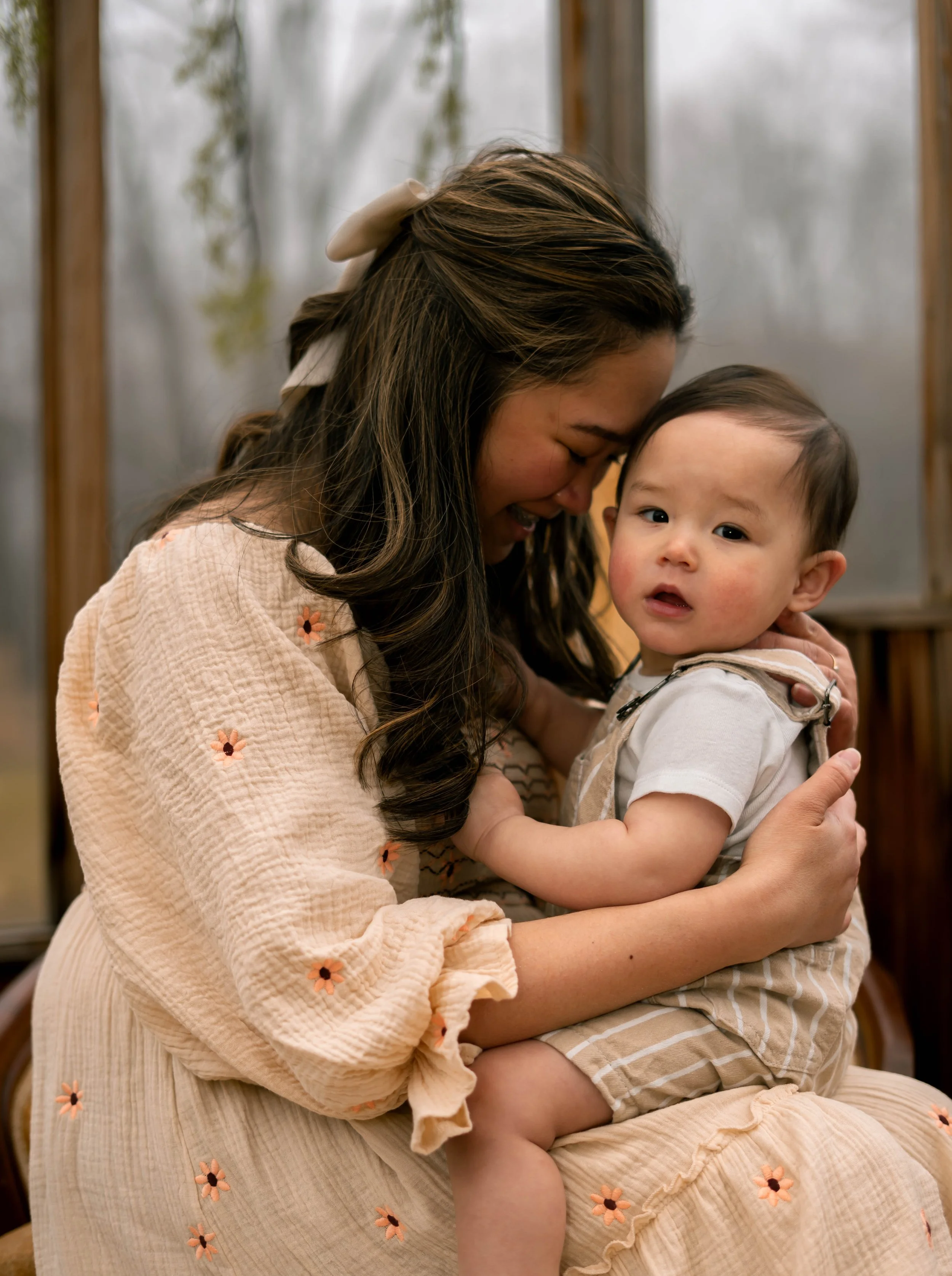 A woman with long brown hair holding a young child with short dark hair, both with their eyes closed, sharing a tender moment indoors with a blurred background.