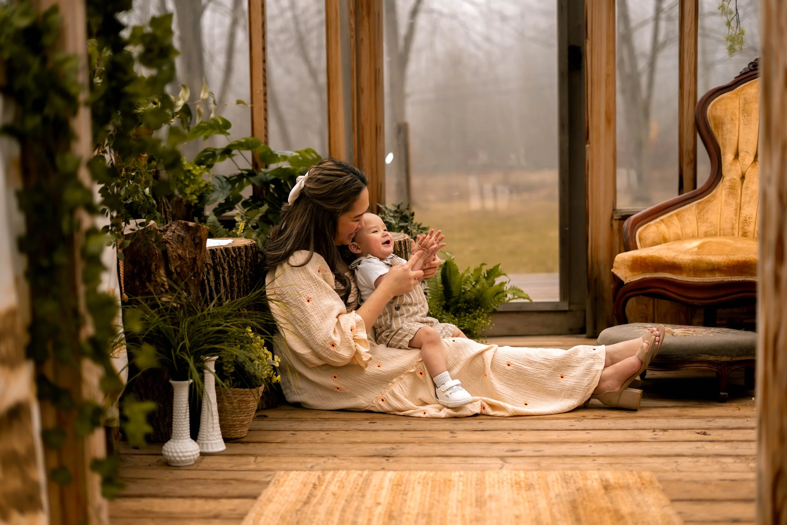 A woman and a young child sitting on a wooden porch, smiling and enjoying each other's company during a foggy day surrounded by greenery and vintage furniture.