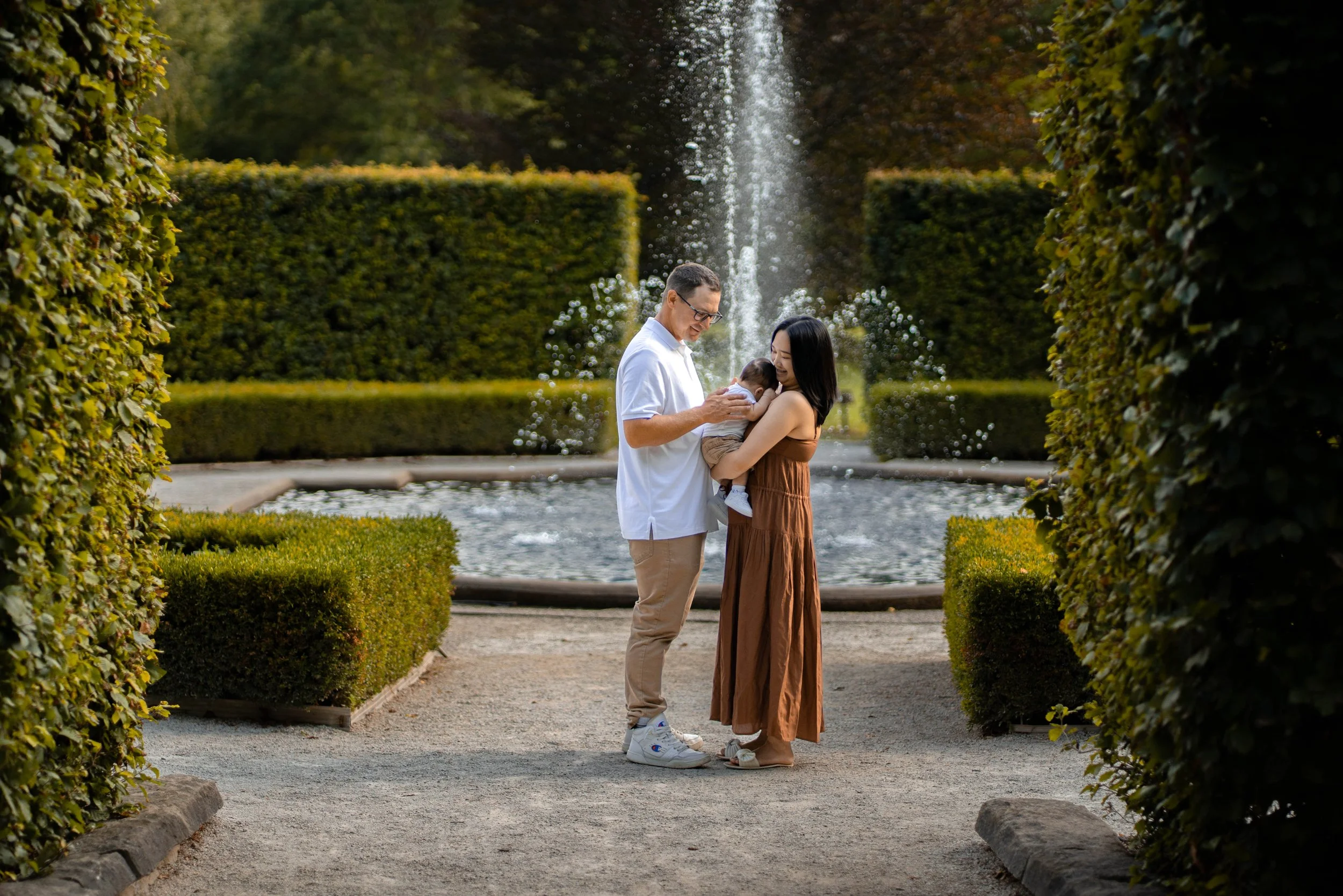 A family of three standing in front of a fountain in a garden. The father is holding a baby, and the mother is standing close to them, all smiling.