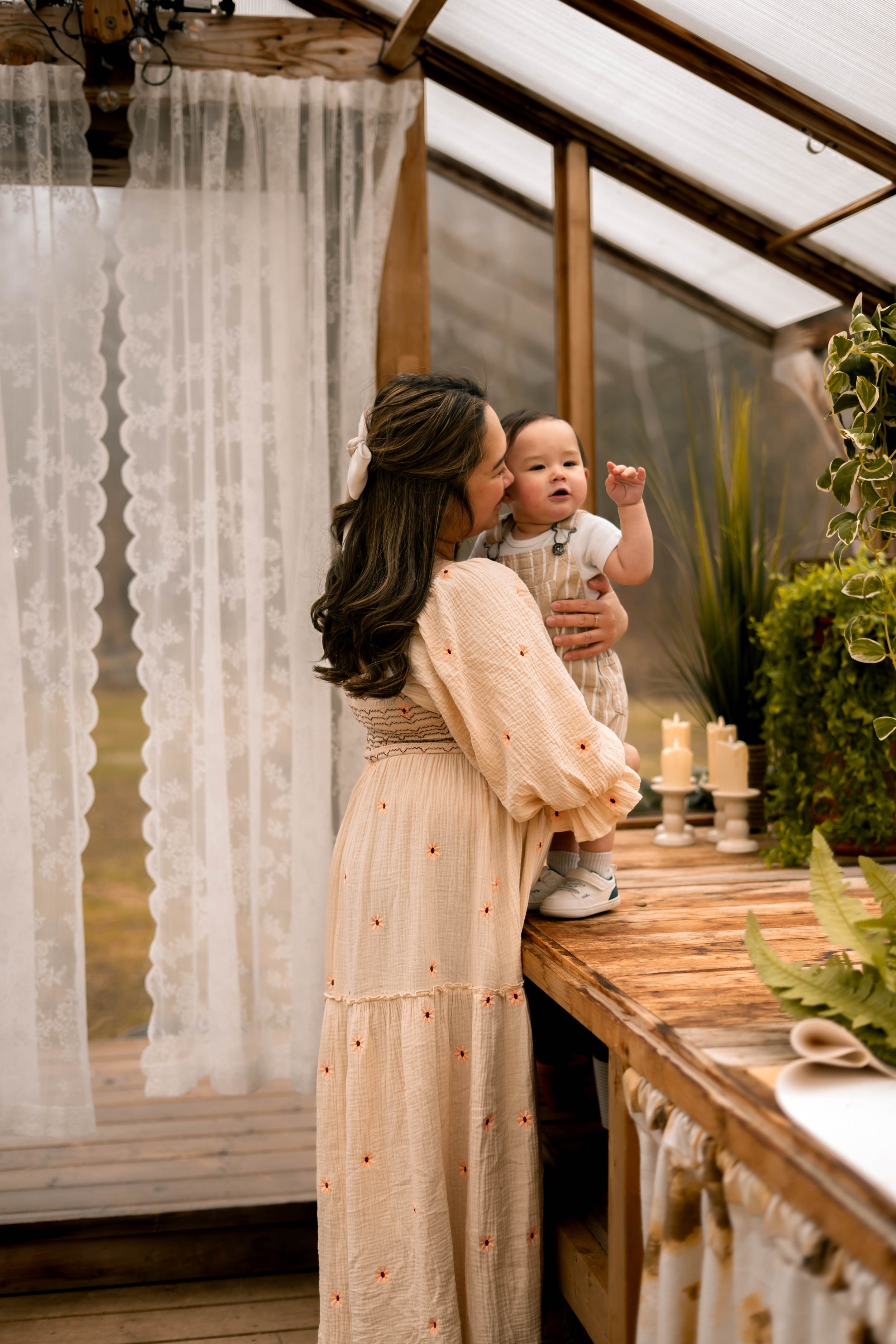 A woman holding a young child inside a rustic greenhouse or plant nursery with lace curtains, wooden beams, and green plants in the background, along with a wooden table displaying candles and fabric.