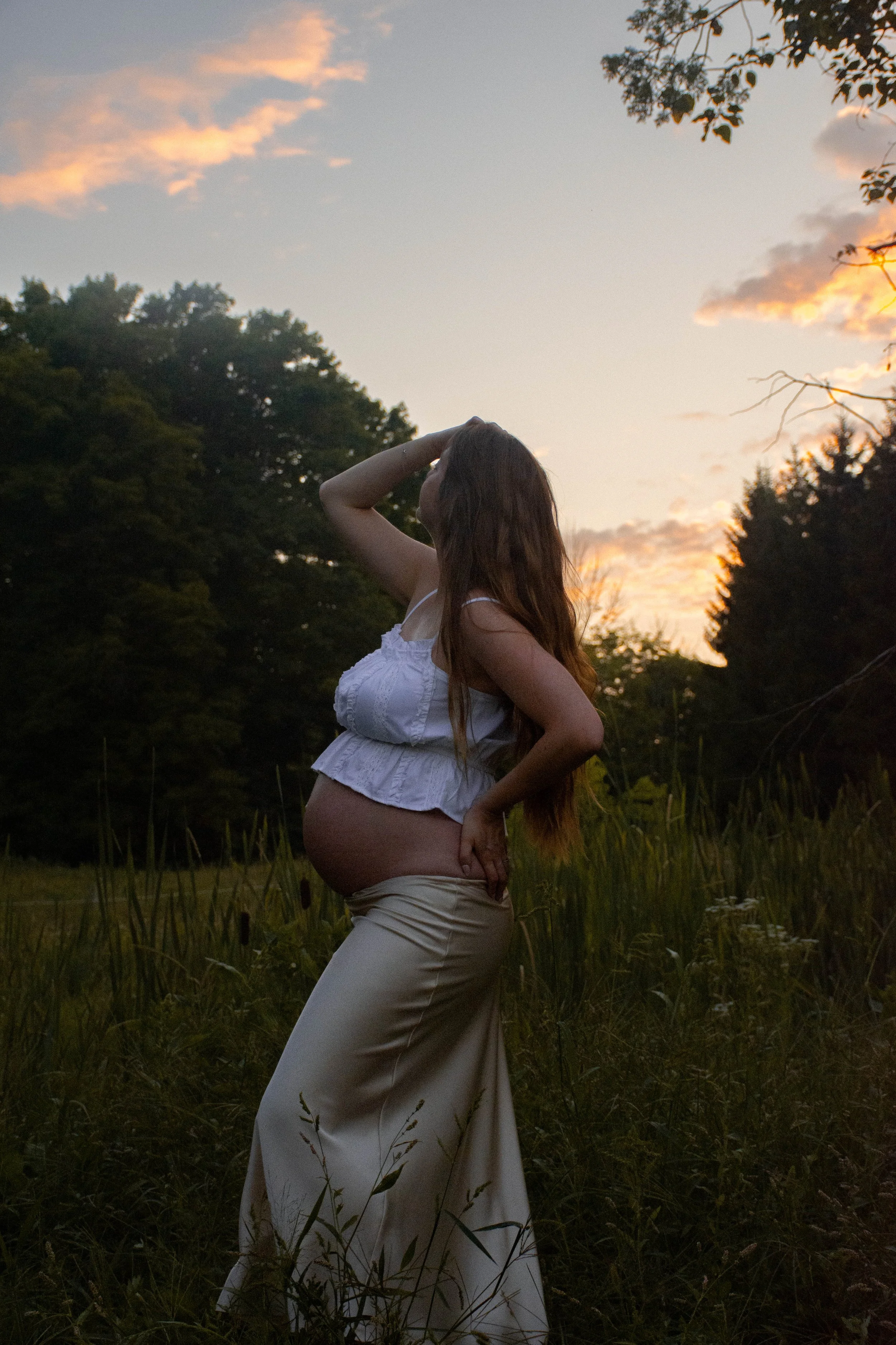 Pregnant woman standing in nature during sunset, wearing a white sleeveless blouse and beige skirt, with one hand on her hip and the other touching her hair.