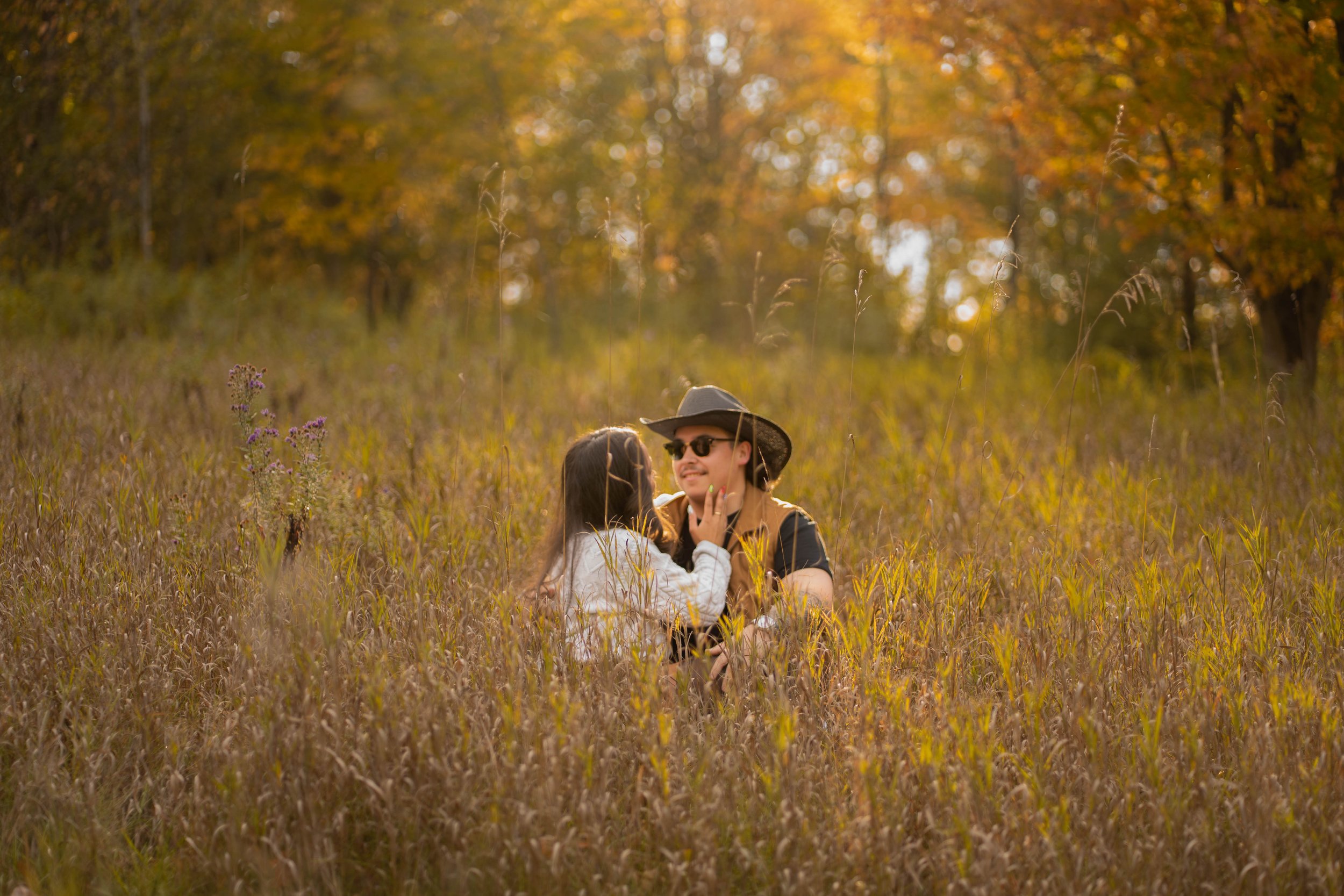 A man and a girl sitting in tall grass in a forested area during autumn, the man wearing sunglasses and a hat, being touched gently by the girl.